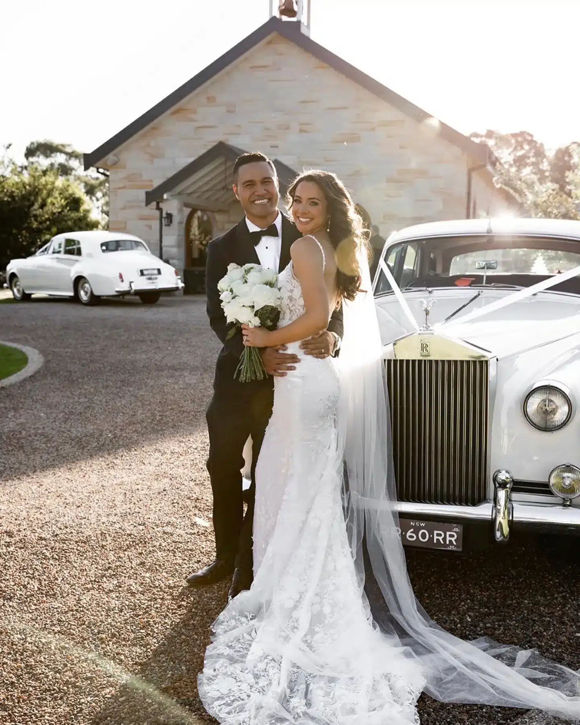 Smiling bride and groom embrace beside a classic white car in front of a stone church at sunset.