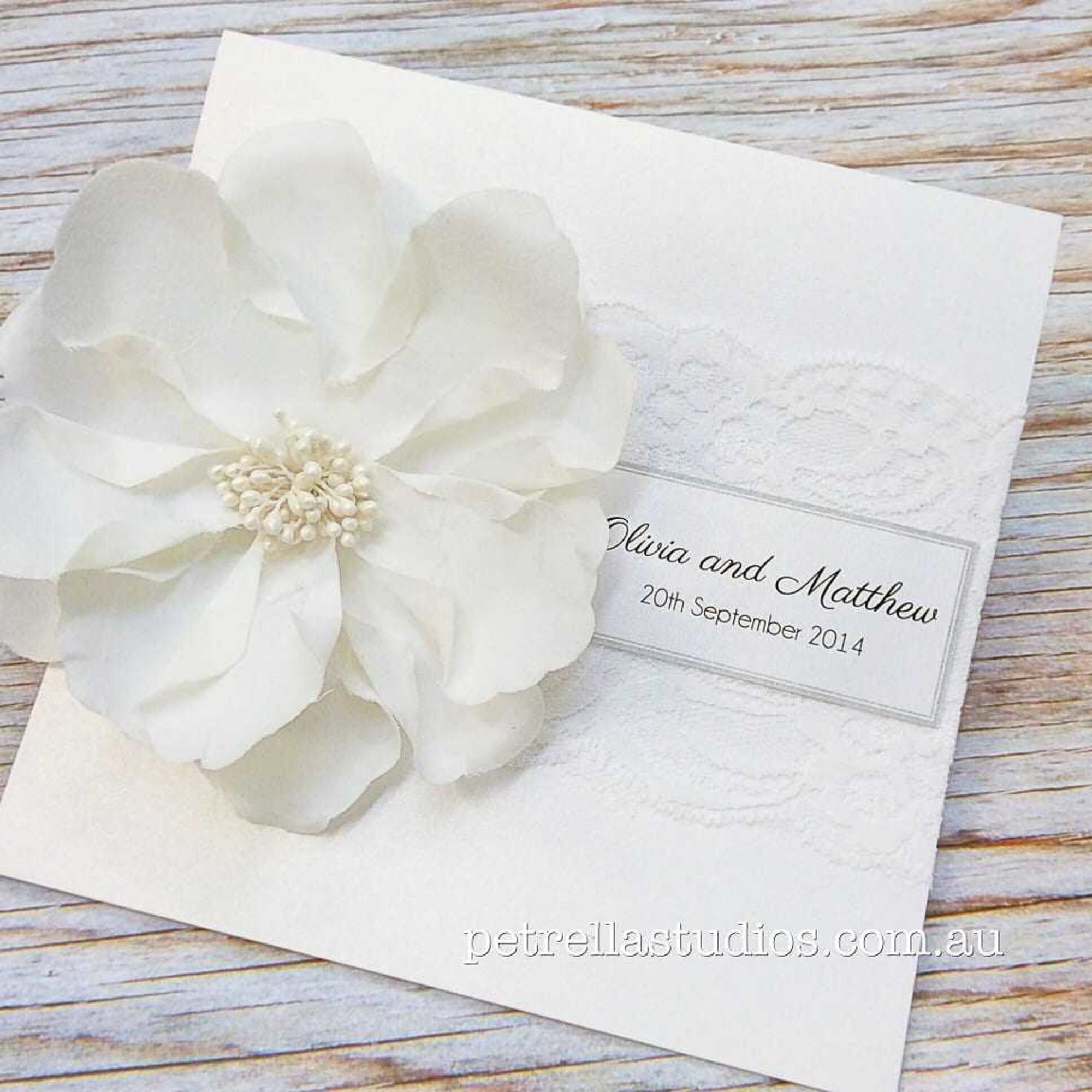 Elegant white wedding invitation with large fabric flower and lace detail on rustic wooden background.