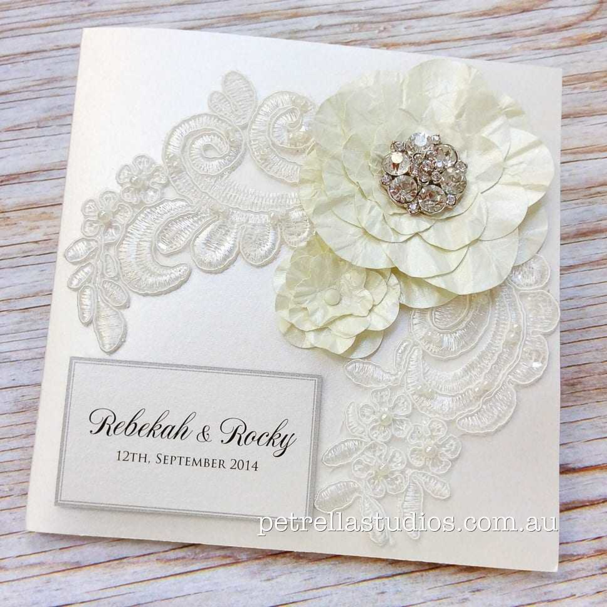 Elegant ivory wedding invitation with lace, layered floral embellishment and crystal accents on a wooden background.