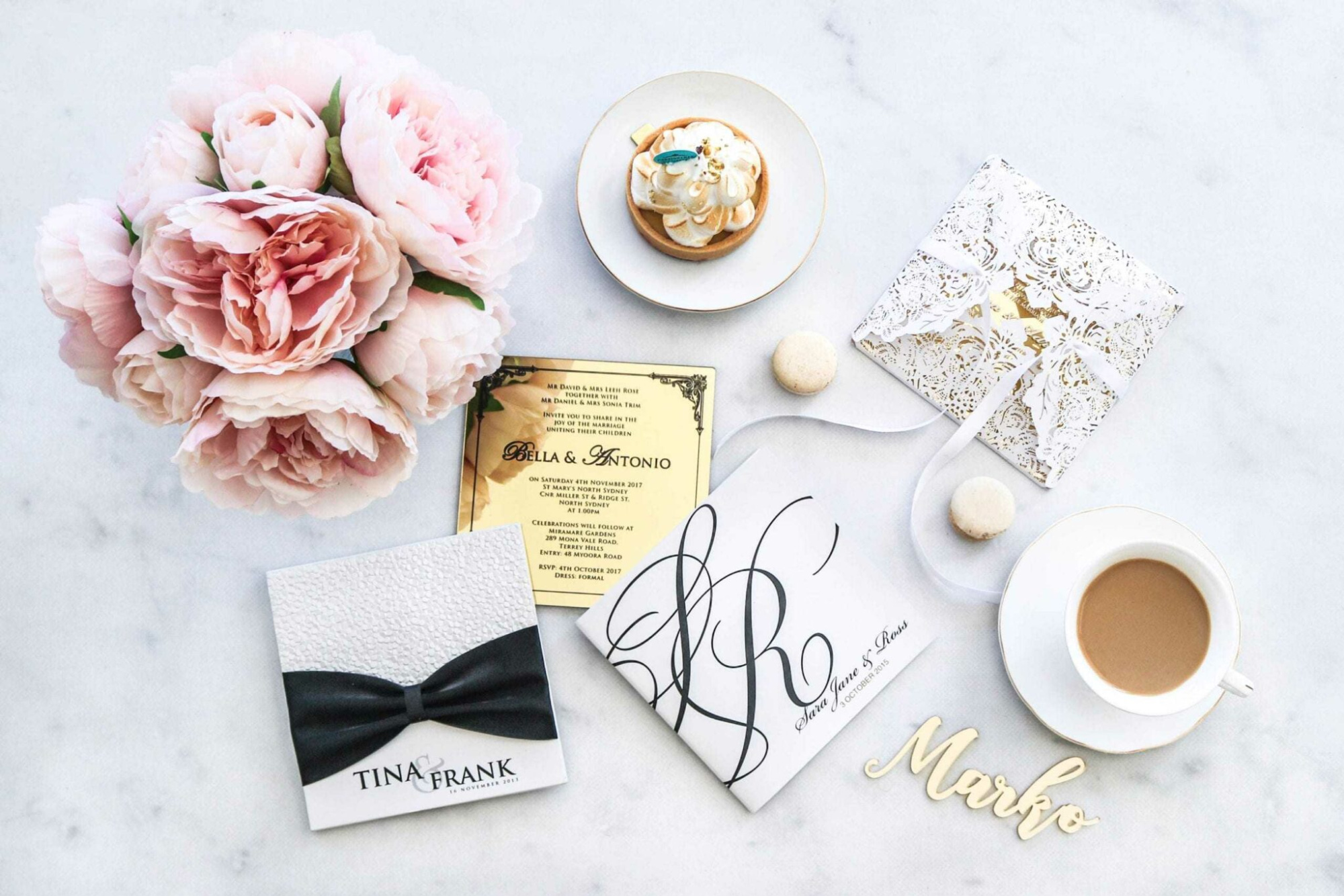 Romantic flatlay of wedding invitations, pastel peonies, dessert, and coffee on a light marble surface.