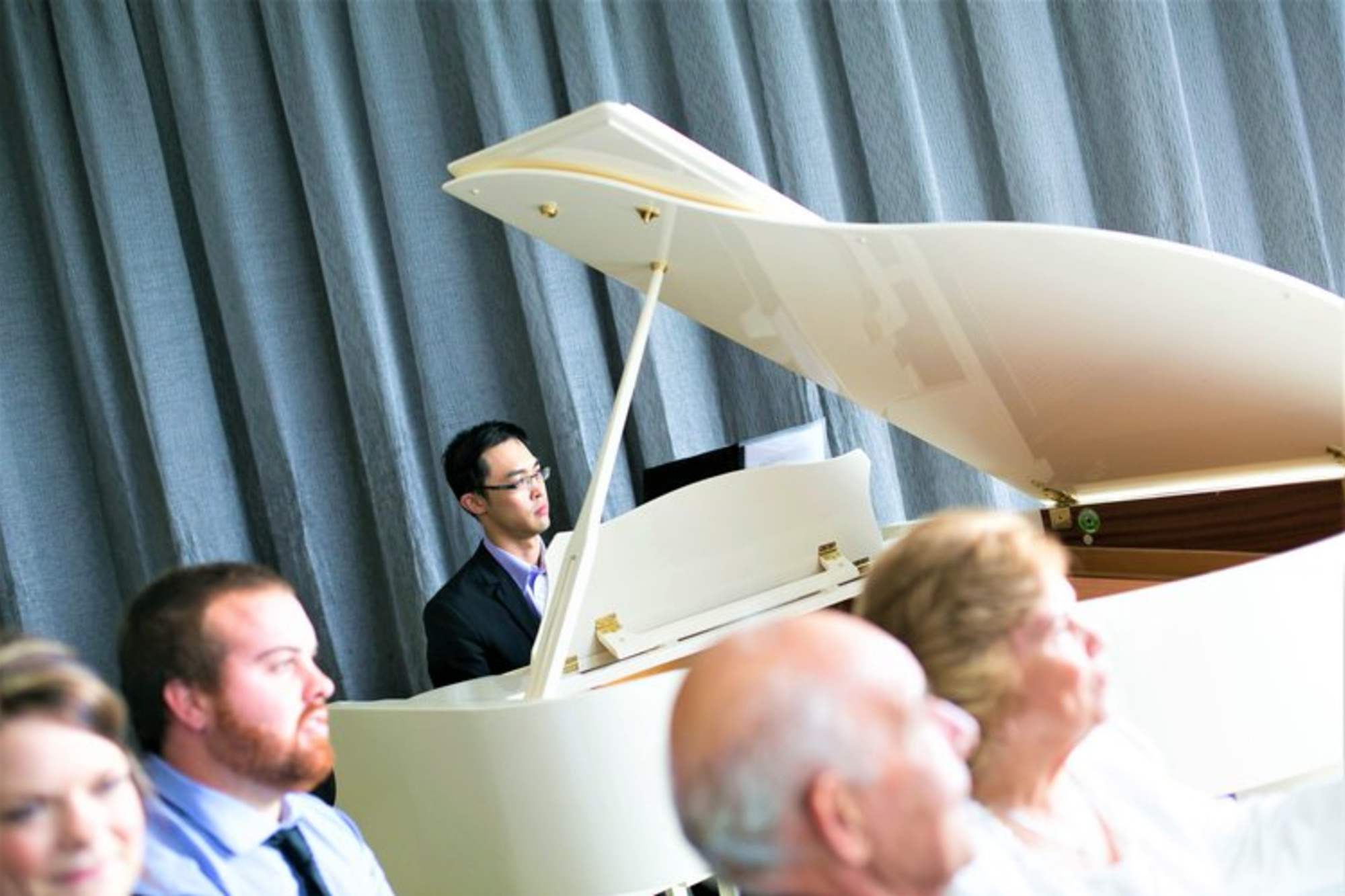 A pianist plays a white grand piano at an indoor wedding ceremony with seated guests in the foreground.