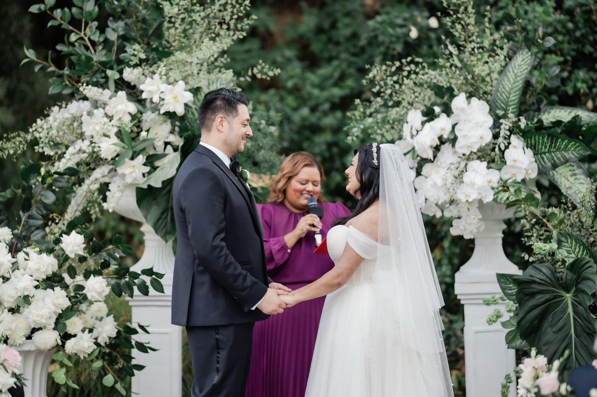 Bride and groom hold hands during an outdoor wedding ceremony framed by lush white florals and greenery.