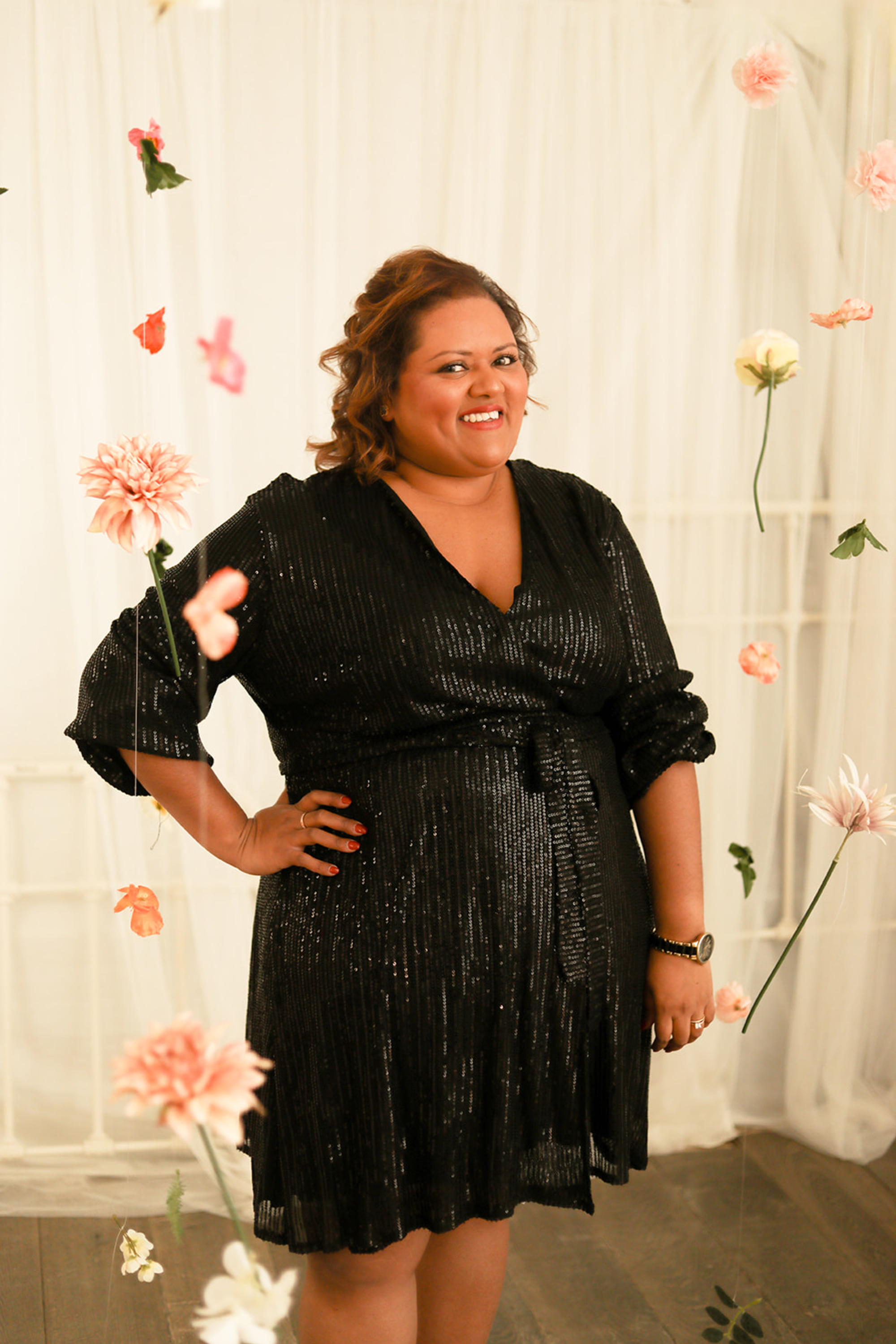 Smiling wedding professional in a black sequin dress poses among hanging pastel flowers on a light backdrop.