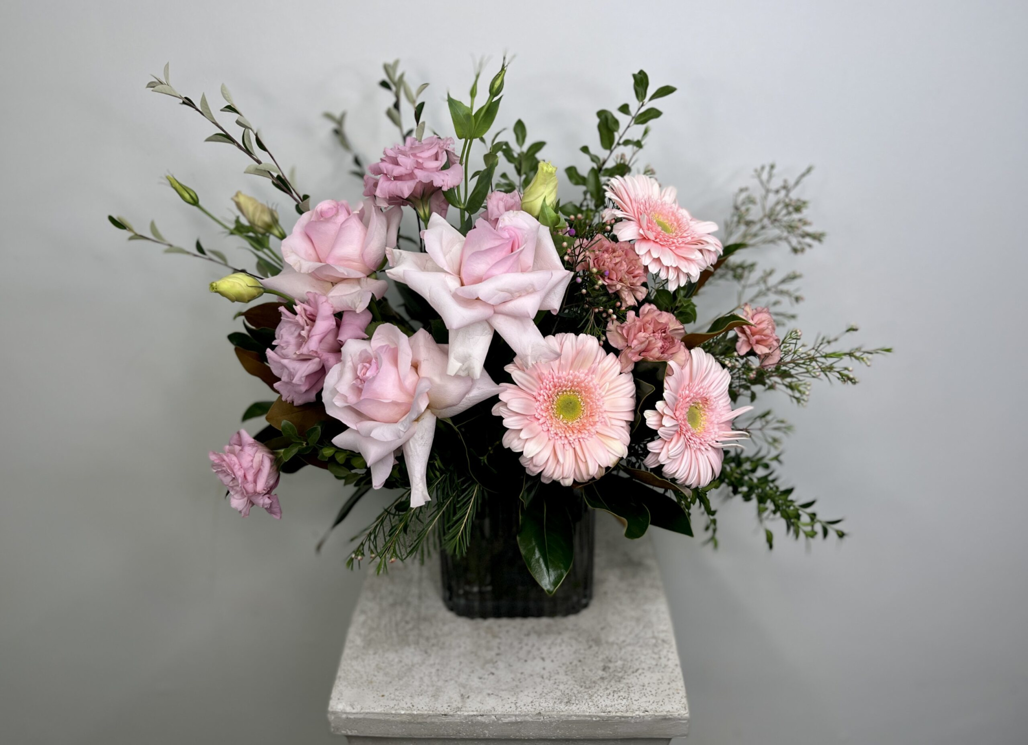 Pink roses and gerbera daisies arranged with greenery in a vase on a stone pedestal.