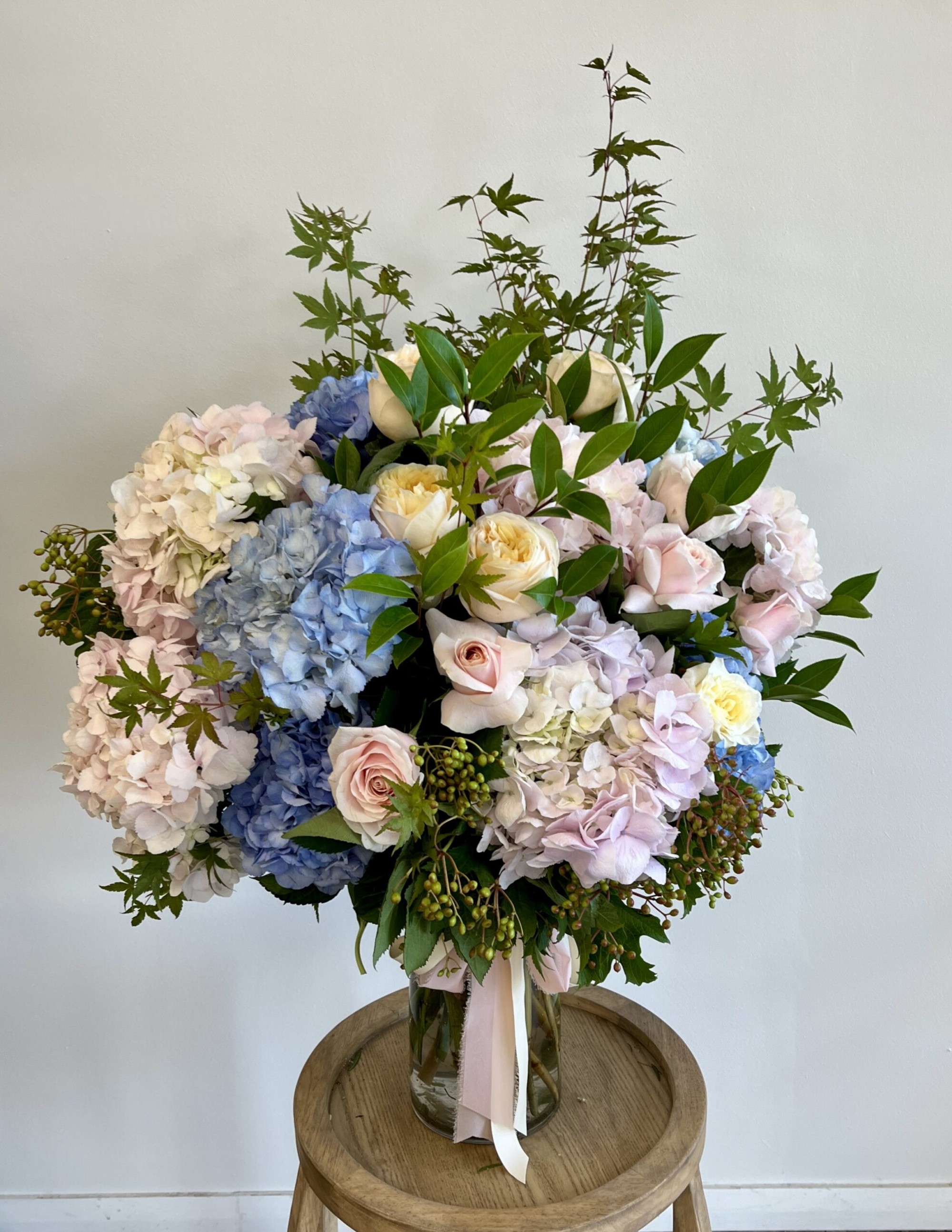 Large pastel floral arrangement with roses and hydrangeas in a glass vase on a wooden stool.