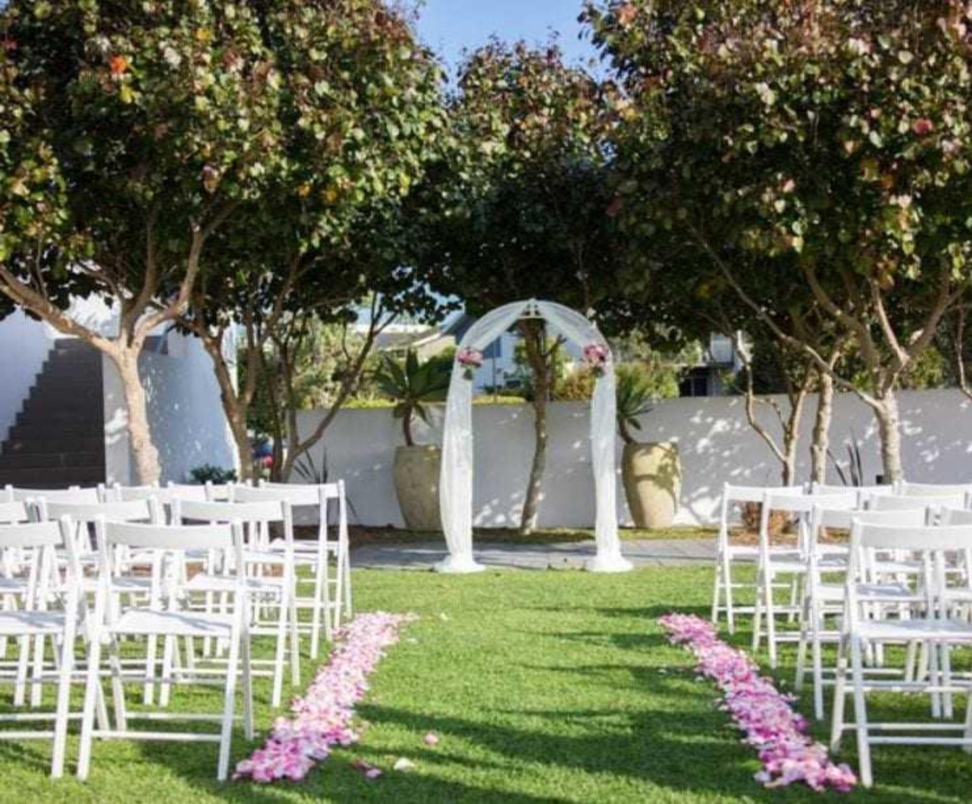 Outdoor wedding ceremony setup with white chairs, a floral arch, and pink rose petals lining the grass aisle.