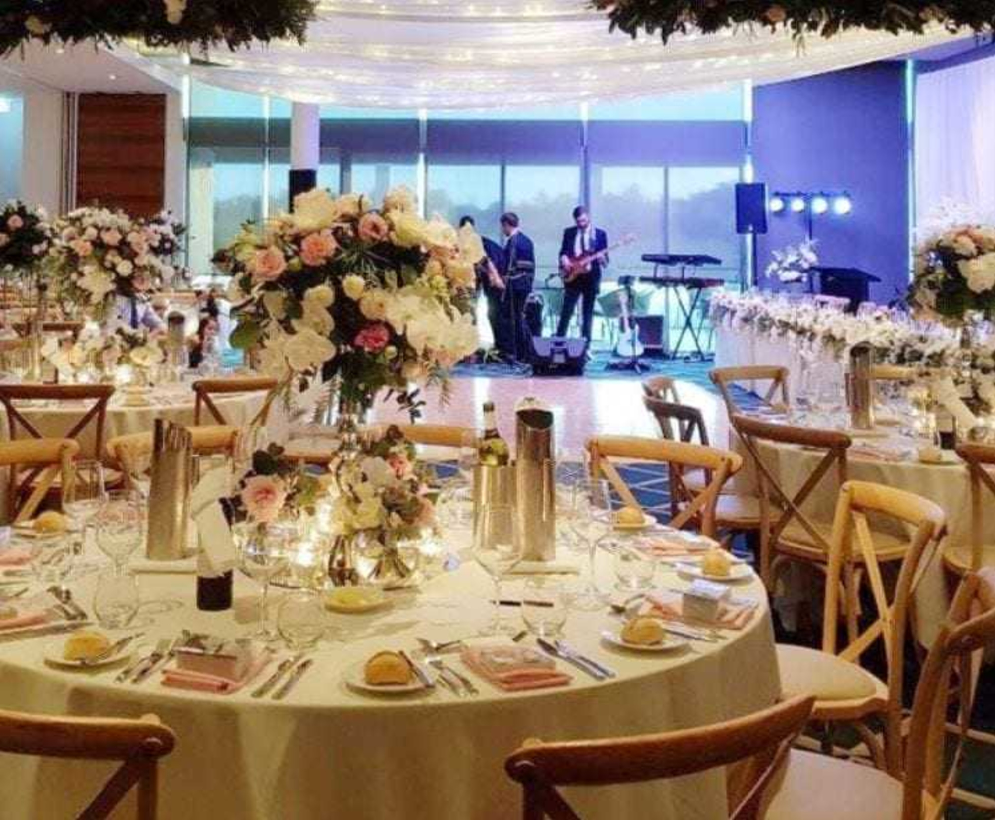 Elegant indoor wedding reception with floral centerpieces, round tables, and a live band on stage.