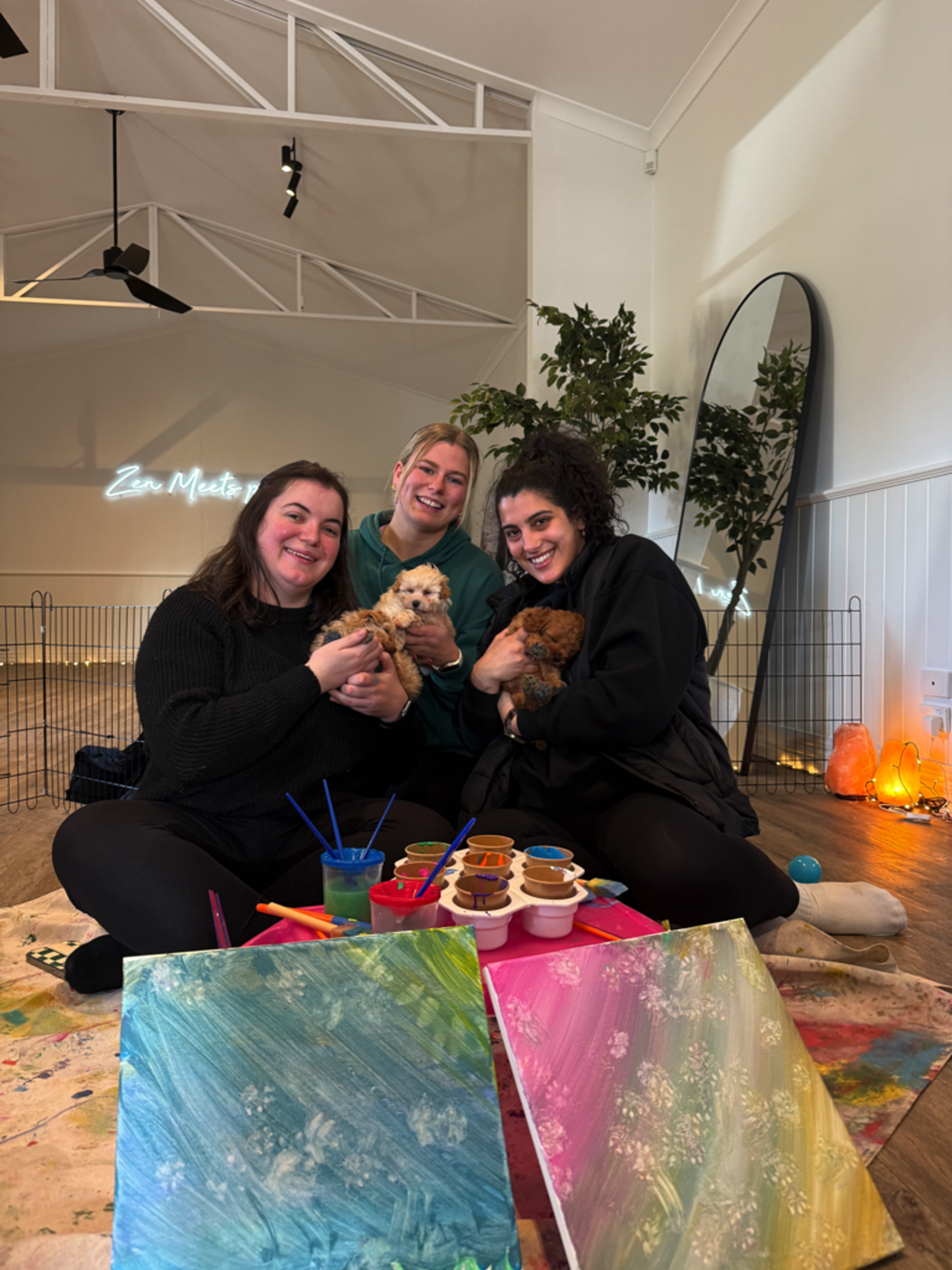 Three women sit on the floor holding small dogs and painting colorful canvases in a bright studio.