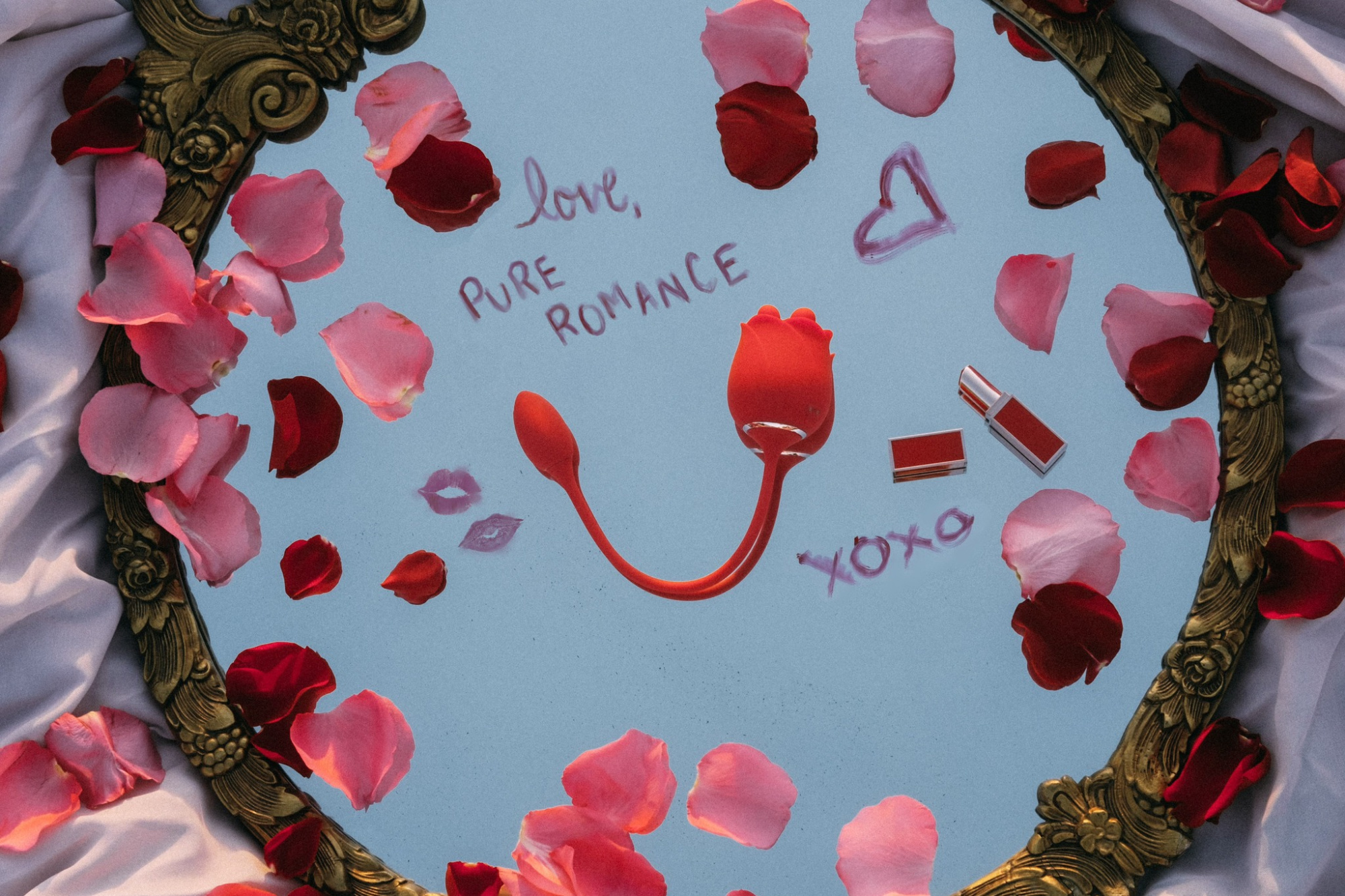 Romantic flatlay of rose petals, love notes, and red accessories arranged on a mirror with a gilded frame.