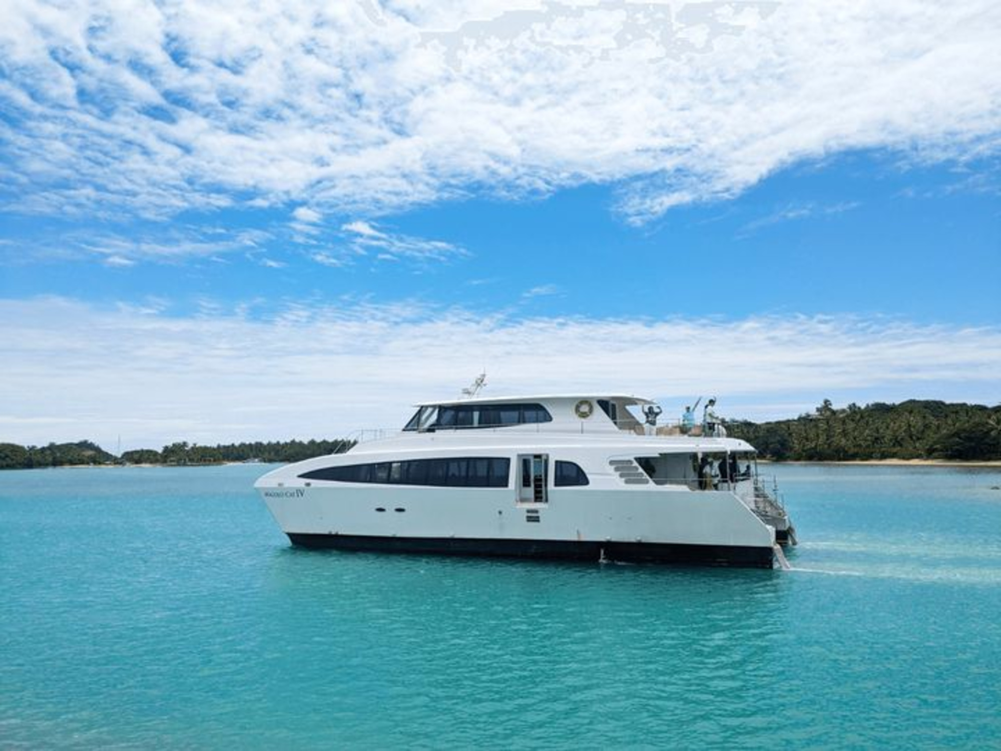 Modern white yacht floating on clear turquoise water near a tropical shoreline under a blue sky with clouds.