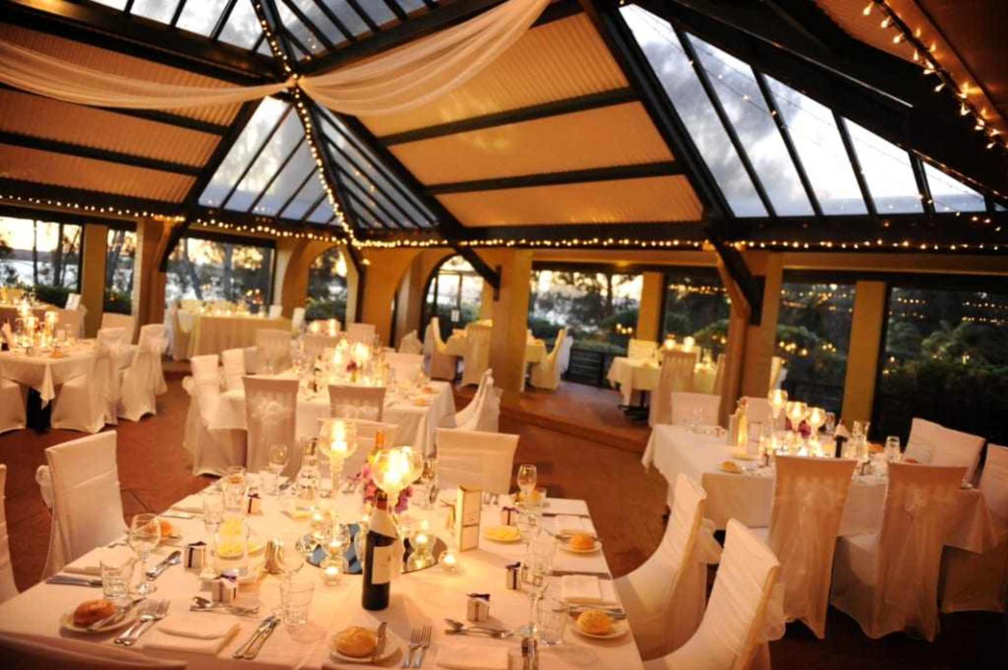 Elegant indoor wedding reception venue with glass ceiling, string lights, and candlelit round tables with white linens.