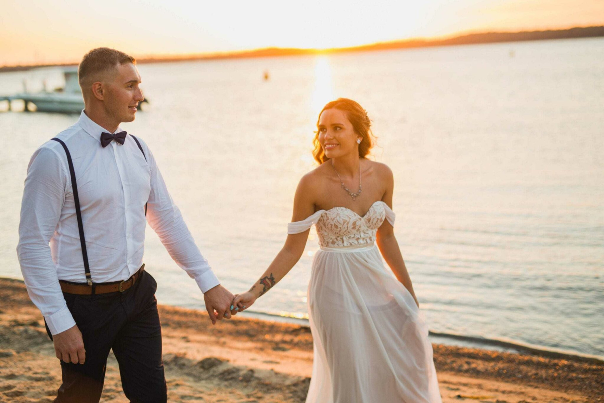 A bride and groom walk hand in hand along the beach at sunset by the water.