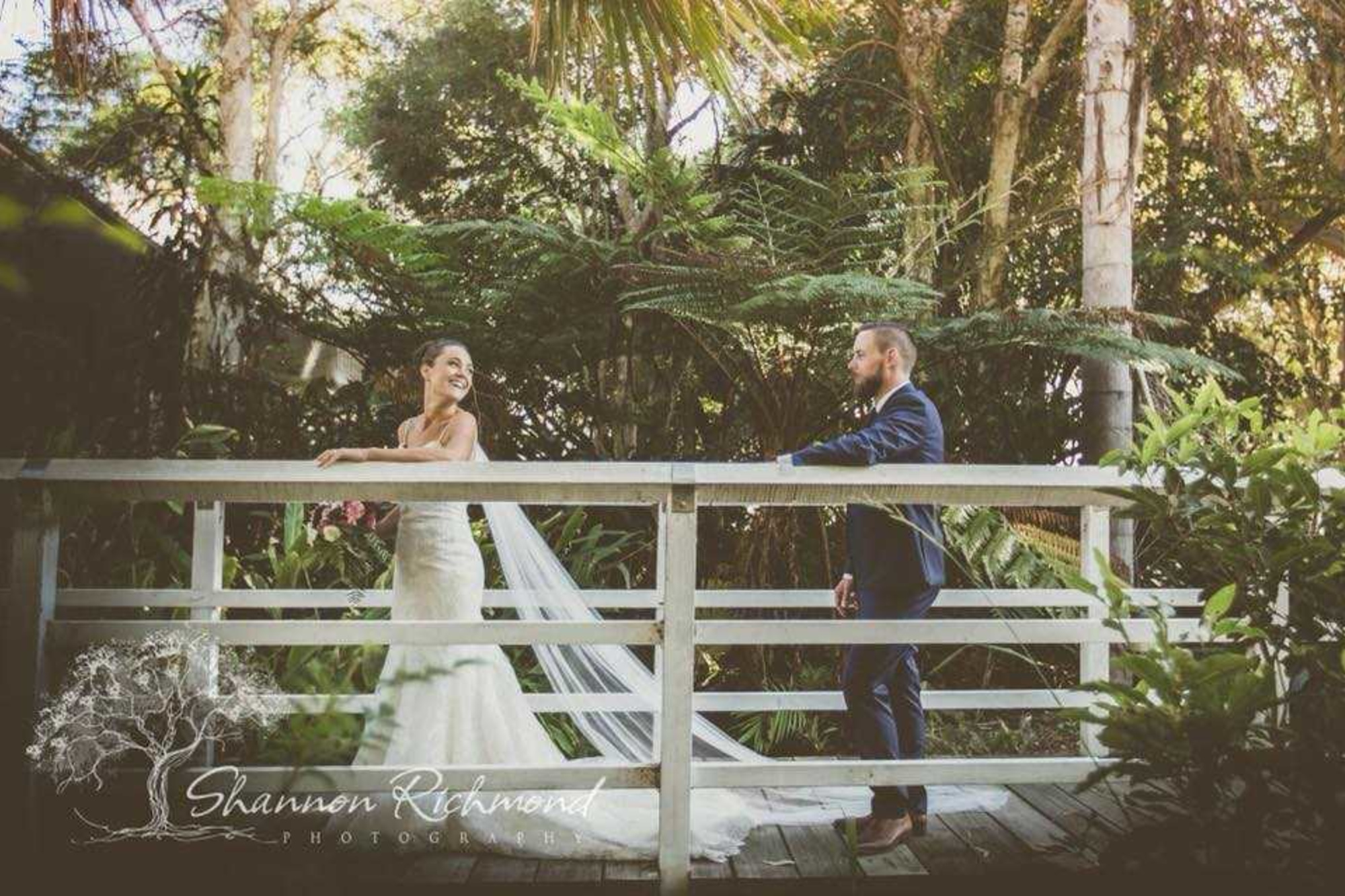 Bride and groom share a moment on a white wooden bridge surrounded by lush tropical greenery.