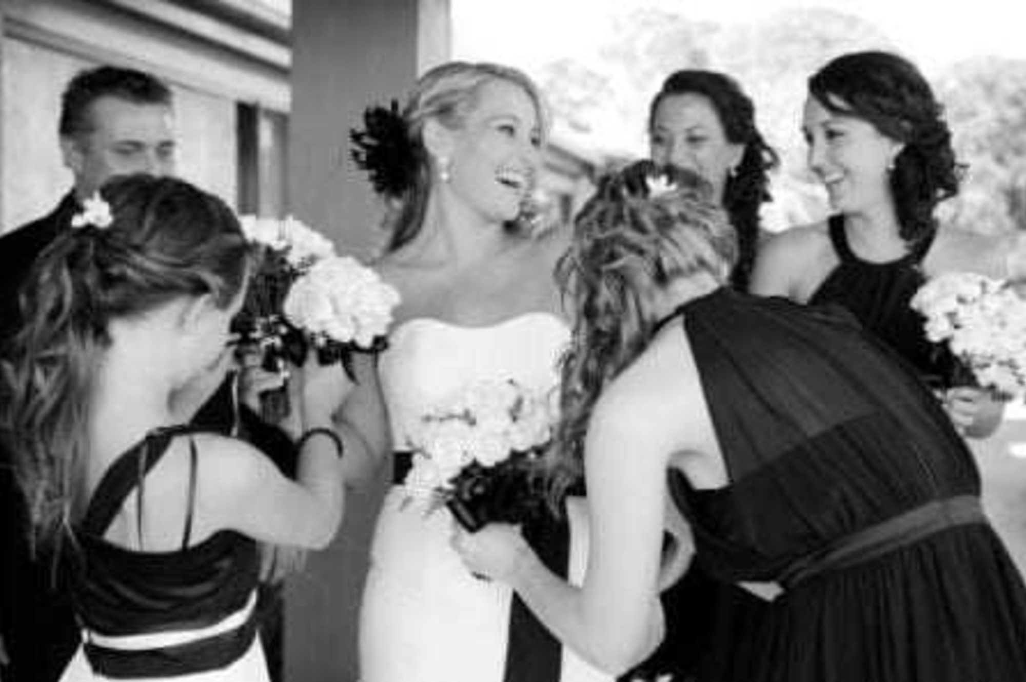 Candid black and white photo of a bride laughing with her bridesmaids as they adjust her dress and hold bouquets.