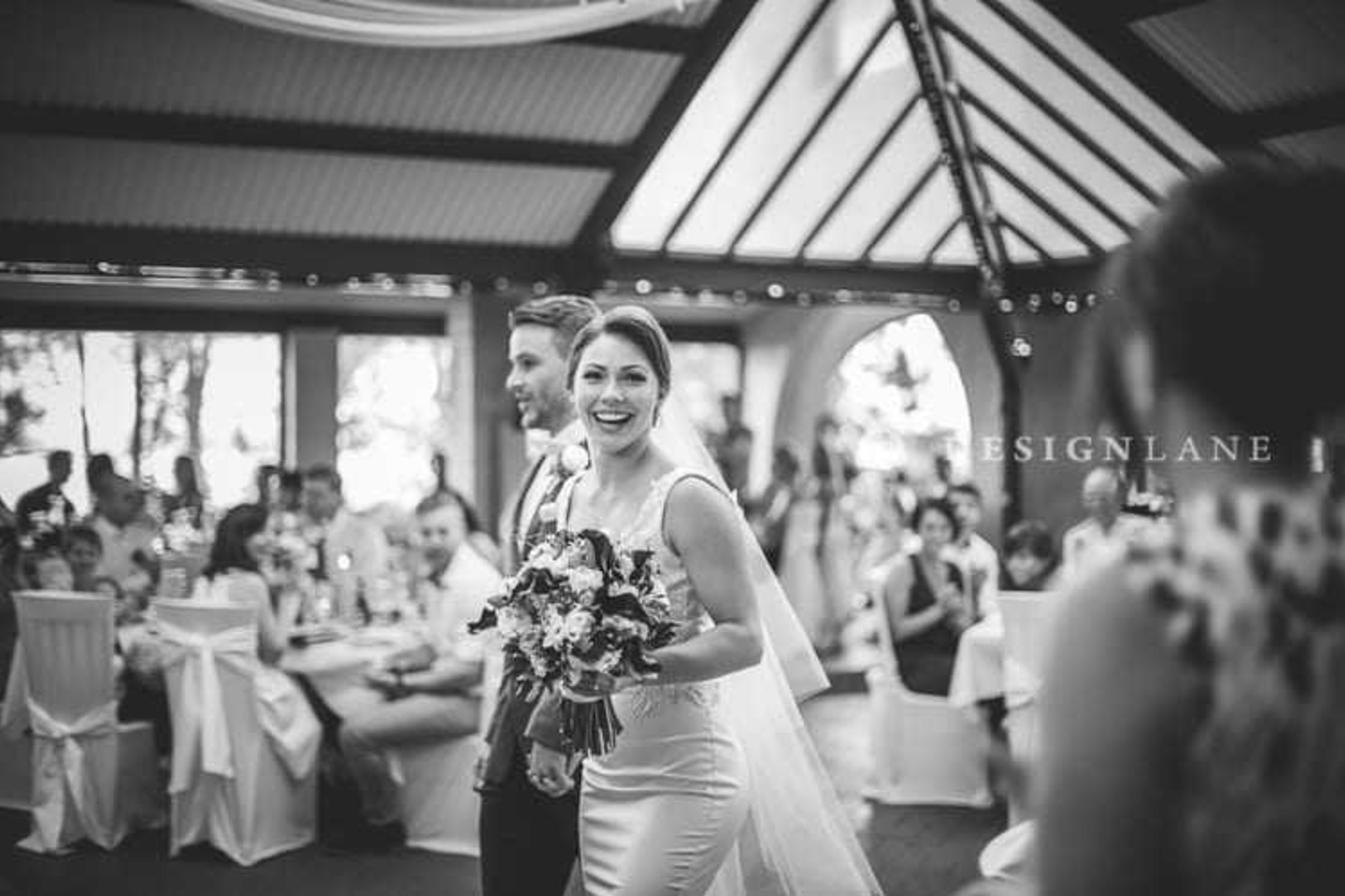 Candid black and white photo of a smiling bride and groom entering their indoor wedding reception among seated guests.