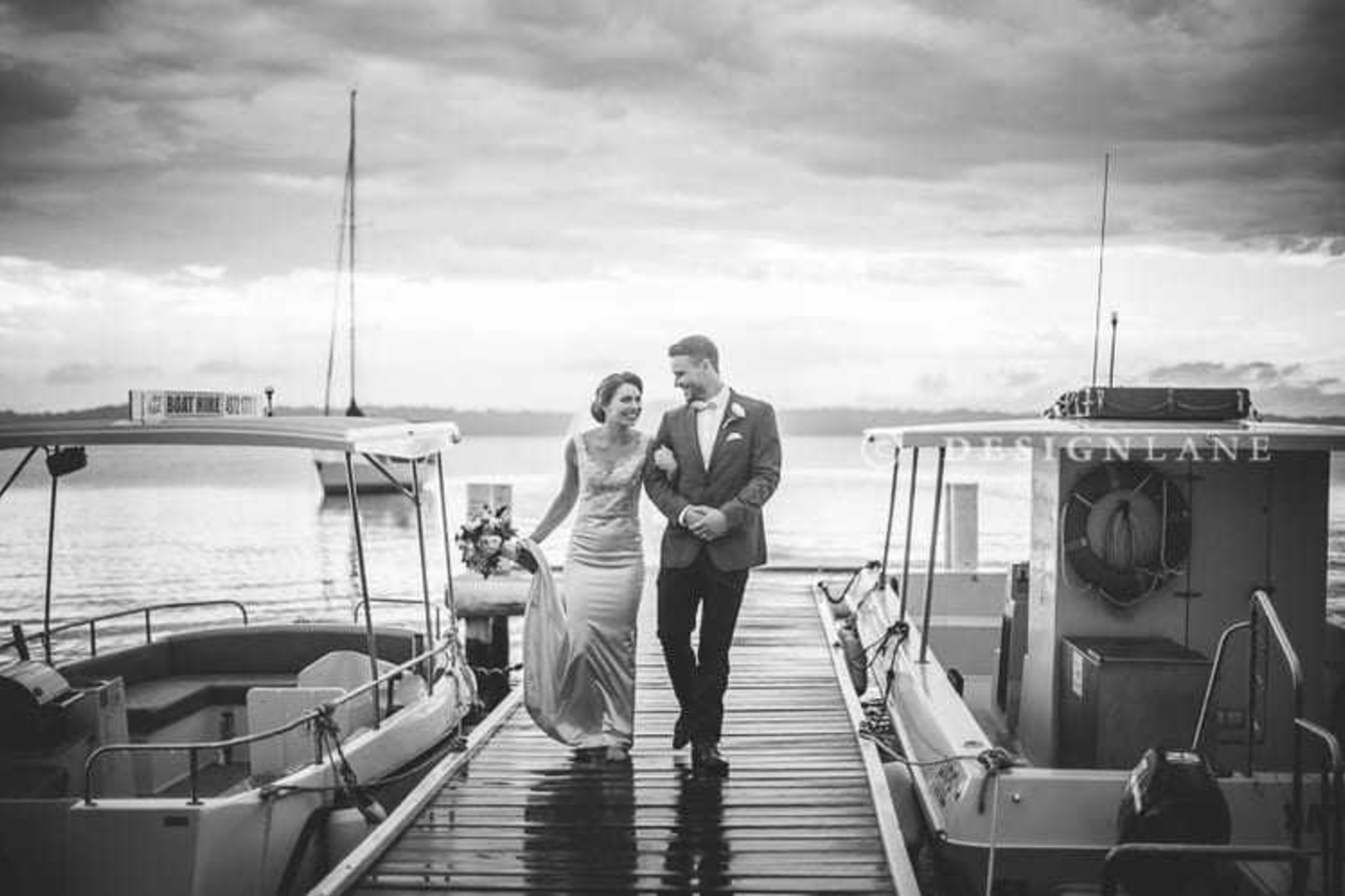 Bride and groom walk arm in arm along a dock between boats on a calm waterfront.