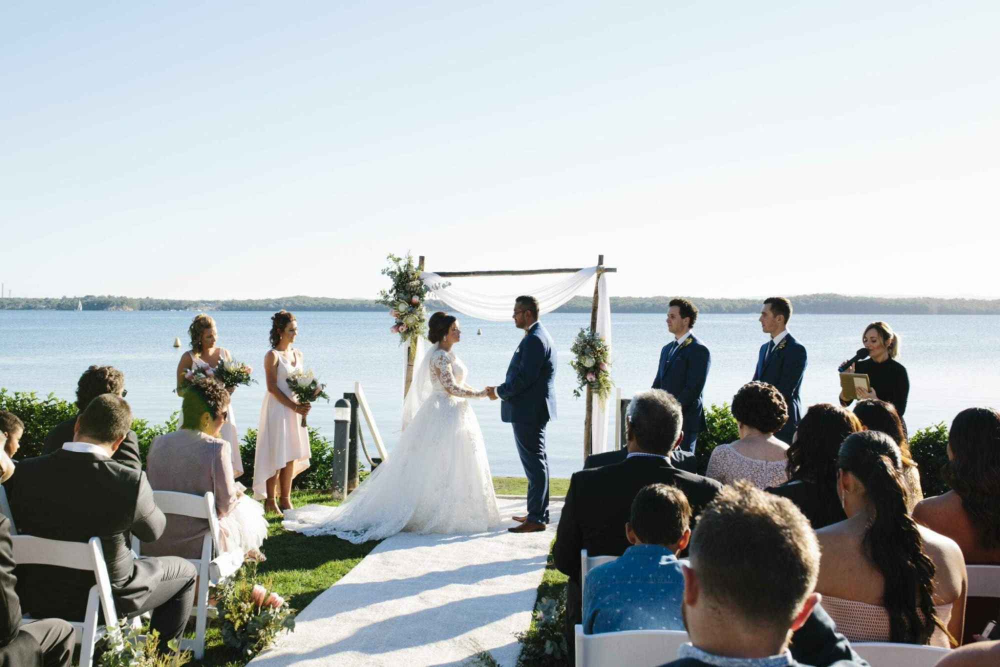 Couple exchanging vows under a floral arch at an outdoor lakeside wedding ceremony with guests seated.