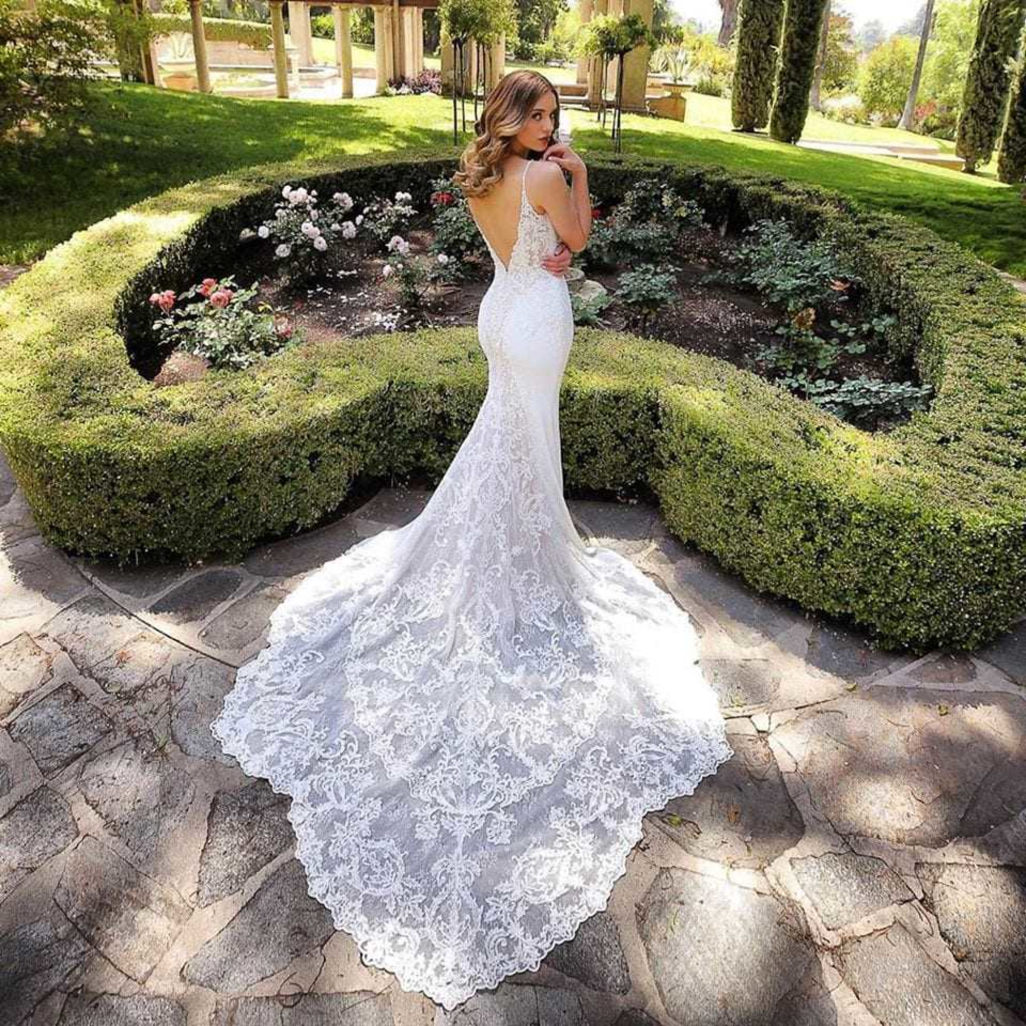 Bride in a backless lace wedding gown with a long train poses in a manicured garden pathway.
