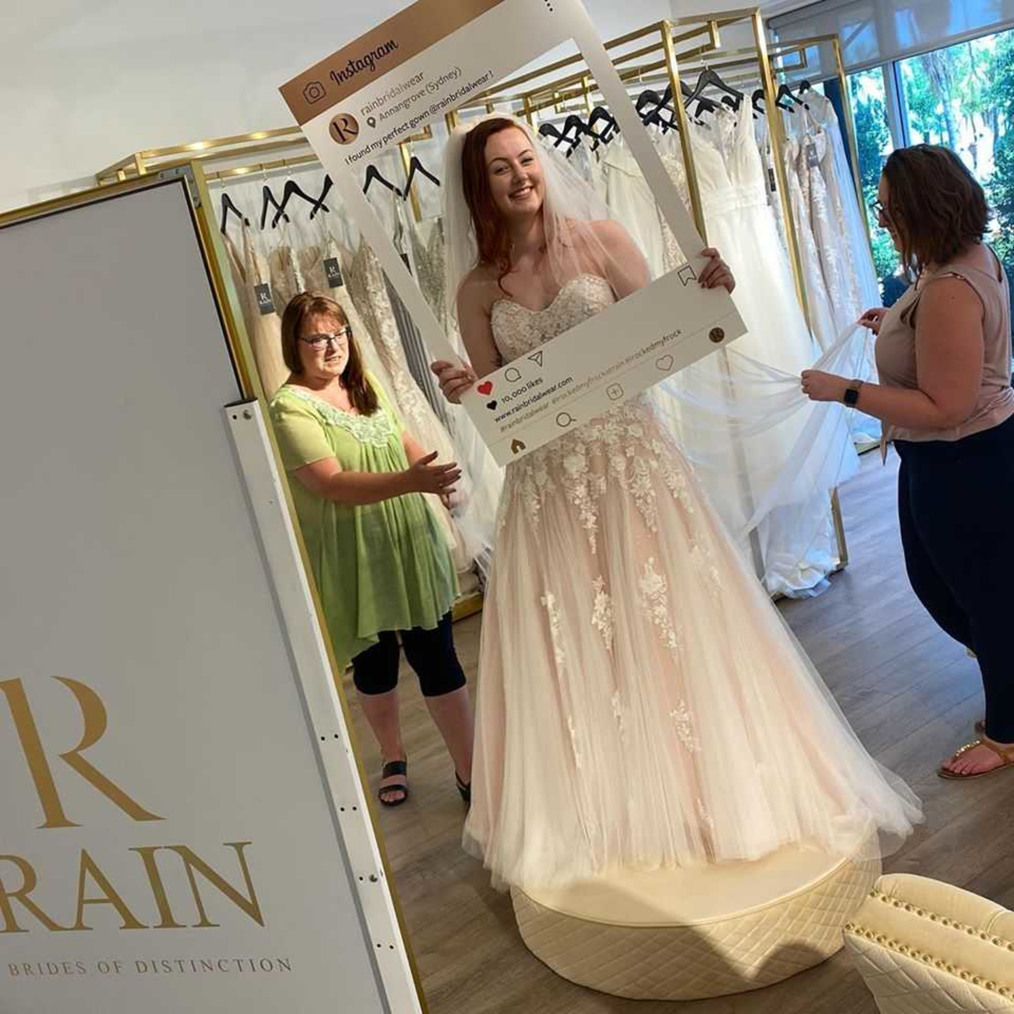 Bride-to-be in a lace tulle gown posing with an Instagram frame during a bridal boutique dress fitting.