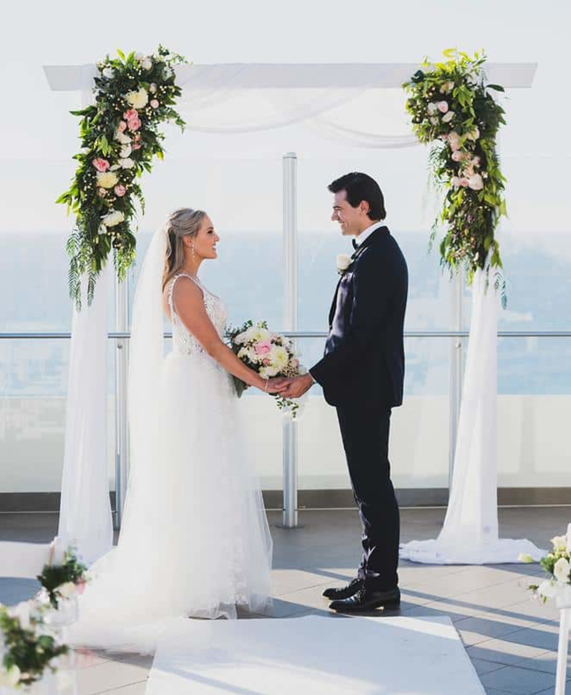 Bride and groom hold hands under a floral arch at a bright seaside rooftop wedding ceremony.