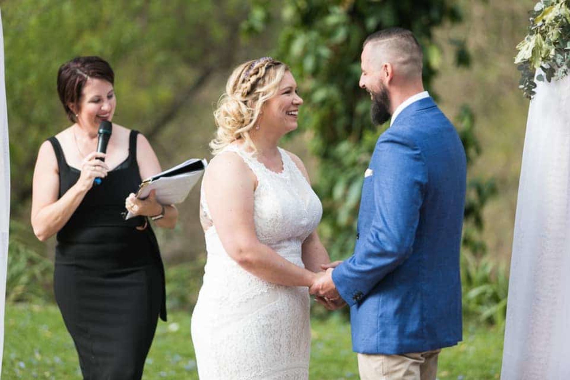 Bride and groom hold hands and smile during an outdoor wedding ceremony with an officiant speaking nearby.