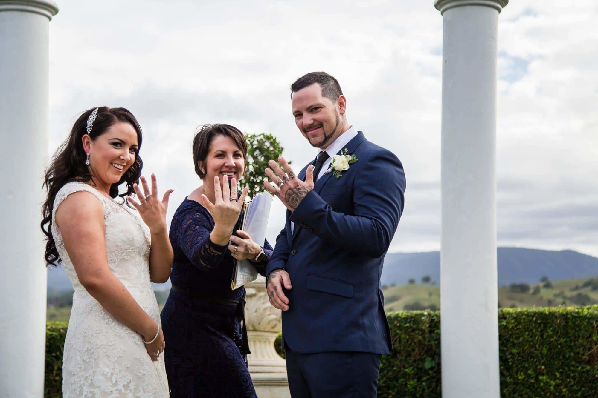 Bride, groom, and celebrant outdoors showing their hands and rings between two white columns with hills in the background.