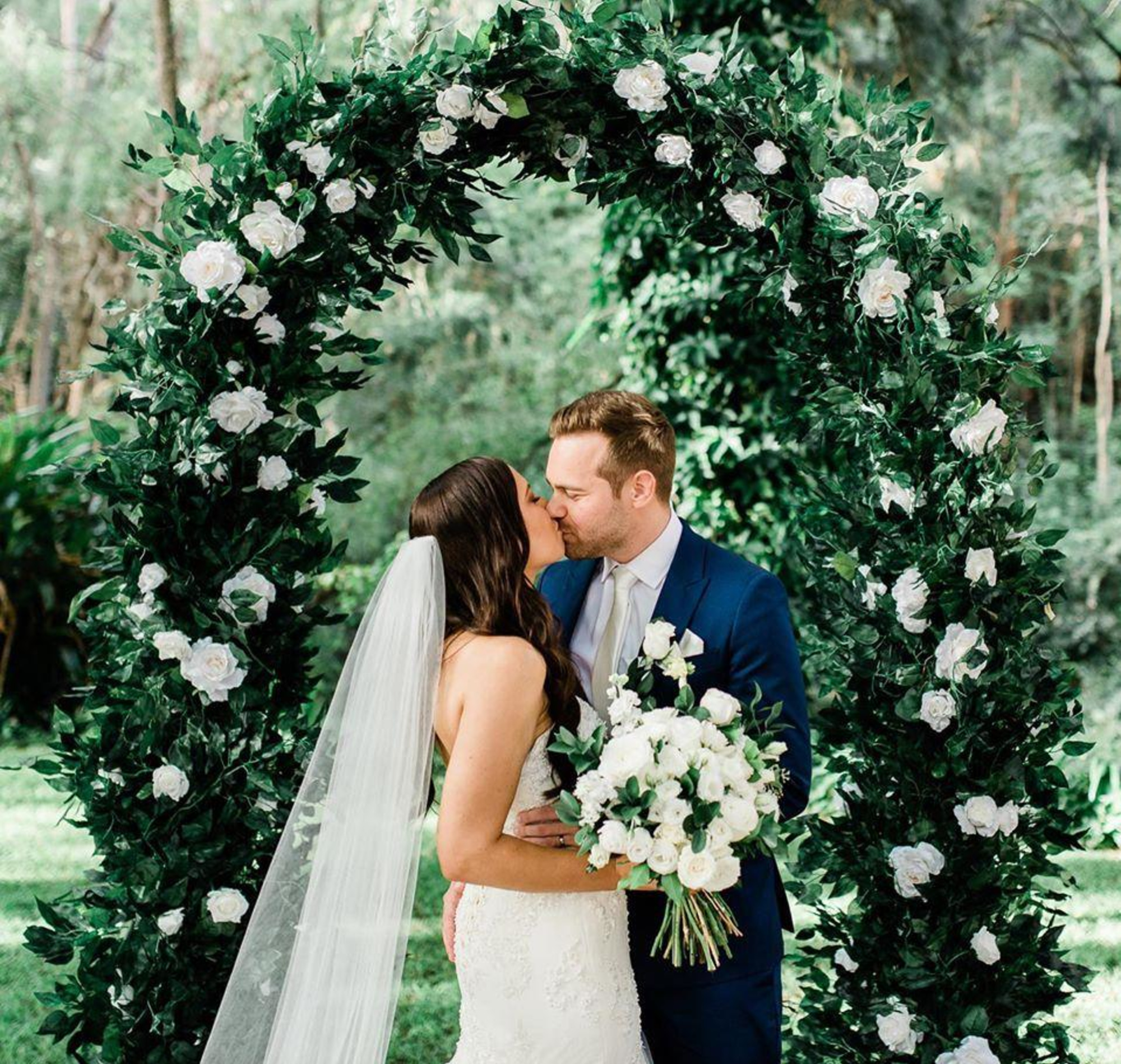 Bride and groom kiss beneath a lush green floral arch in an outdoor garden ceremony.