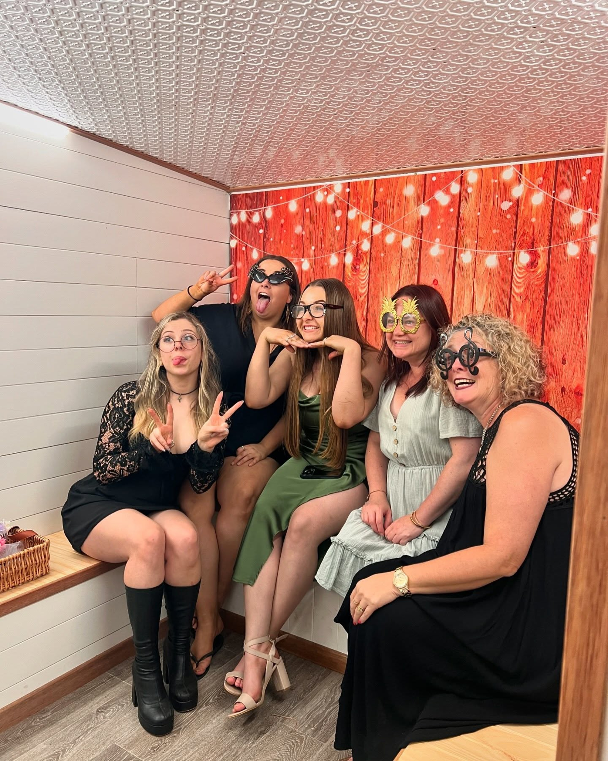 Group of women posing with fun glasses and faces in a wedding photo booth with wood backdrop and string lights.