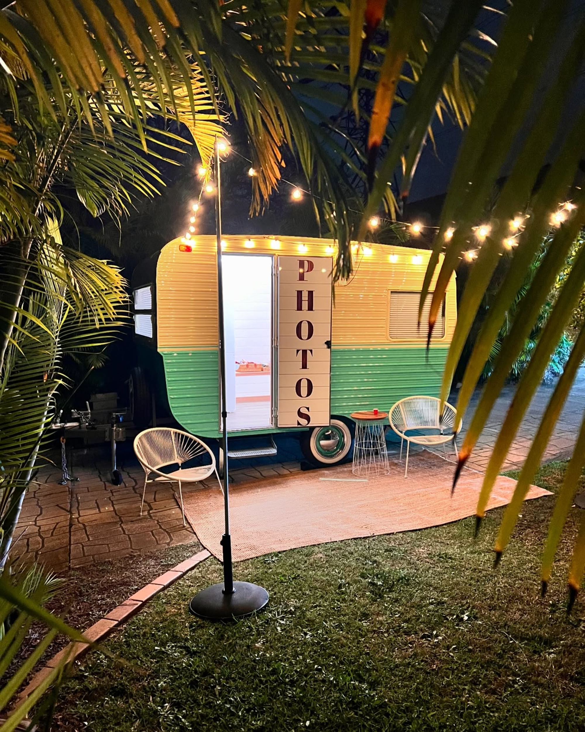 Vintage trailer photo booth set up outdoors at night with string lights and lounge chairs.