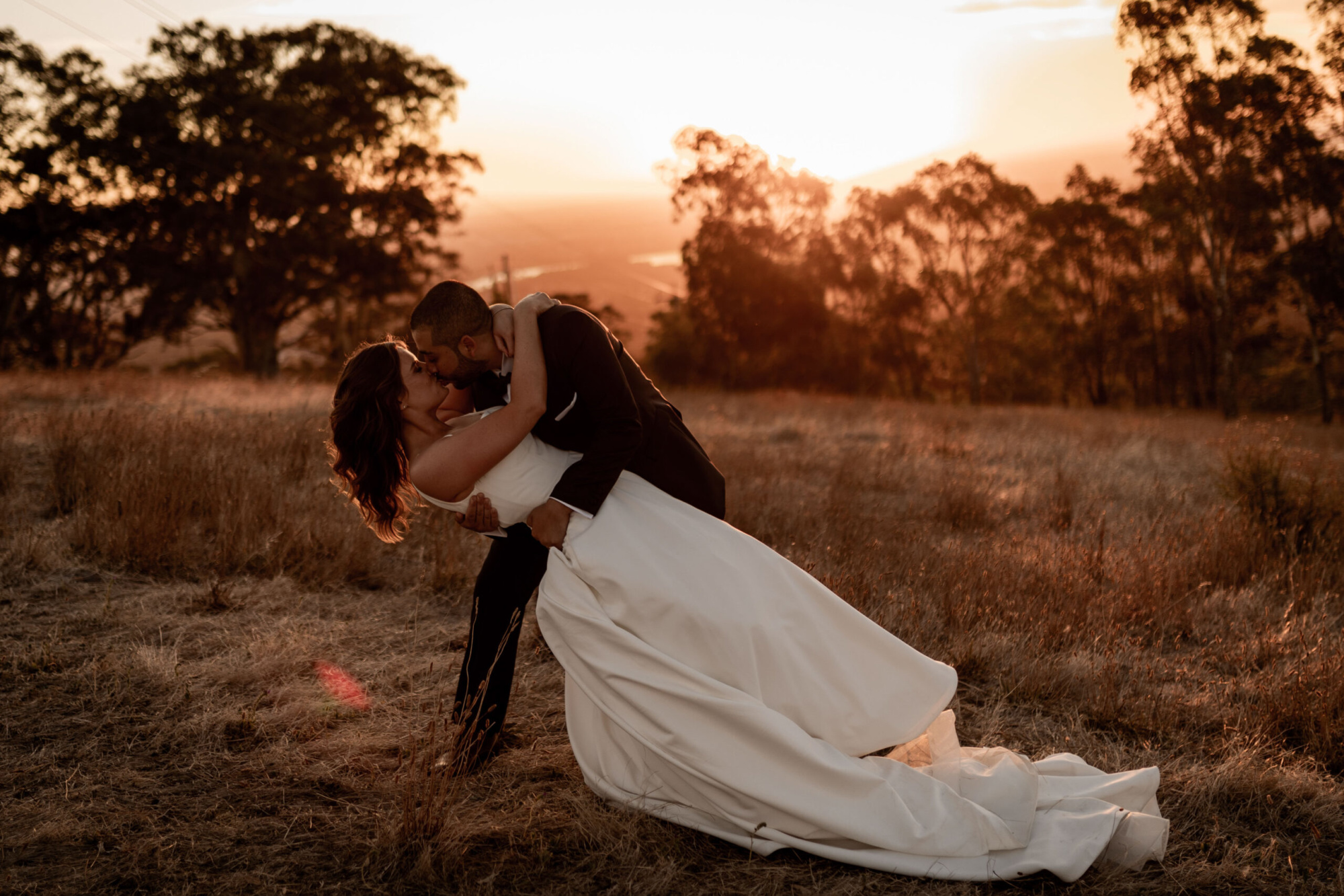 Bride and groom share a romantic dip and kiss in a golden field at sunset.