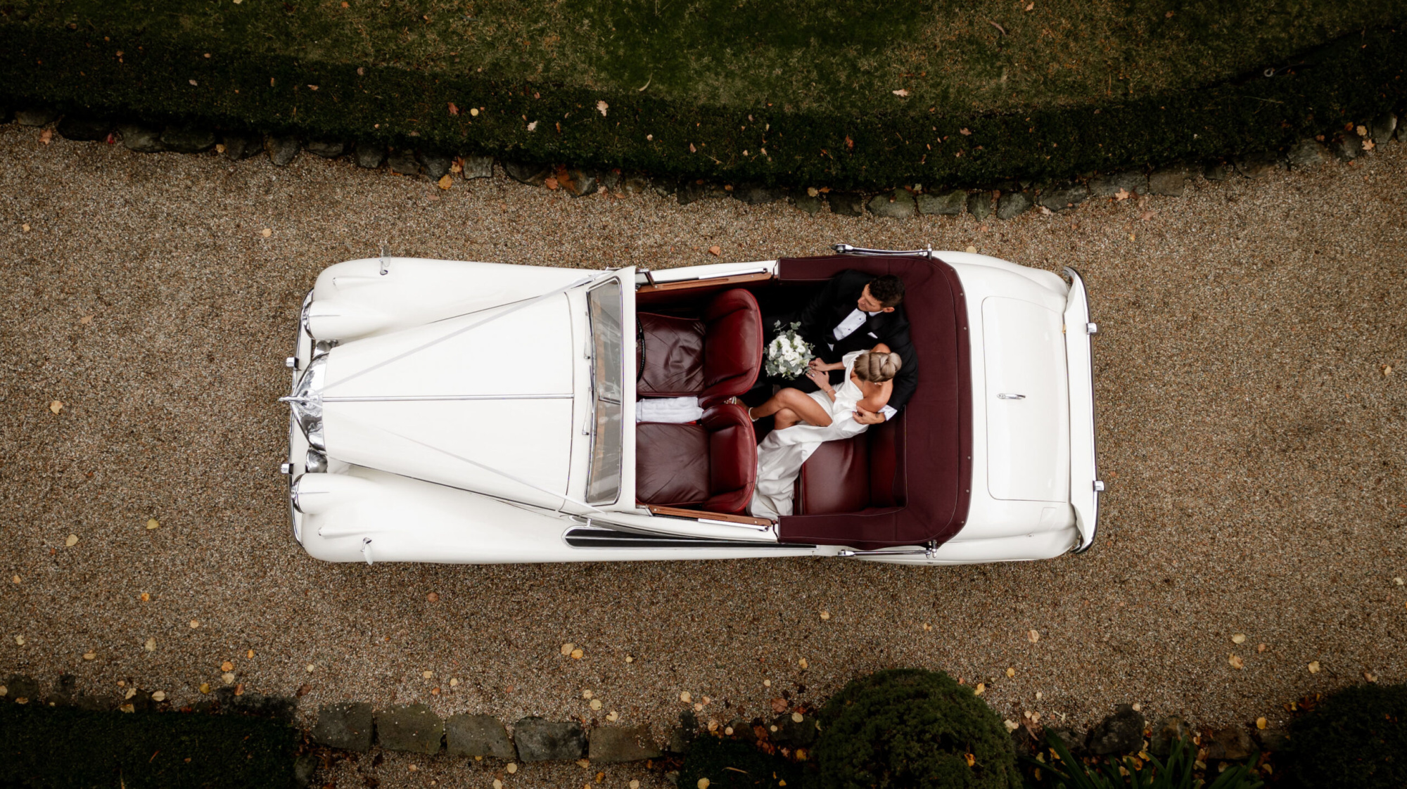A bride and groom sit in a classic white convertible wedding car captured from above on a gravel driveway.