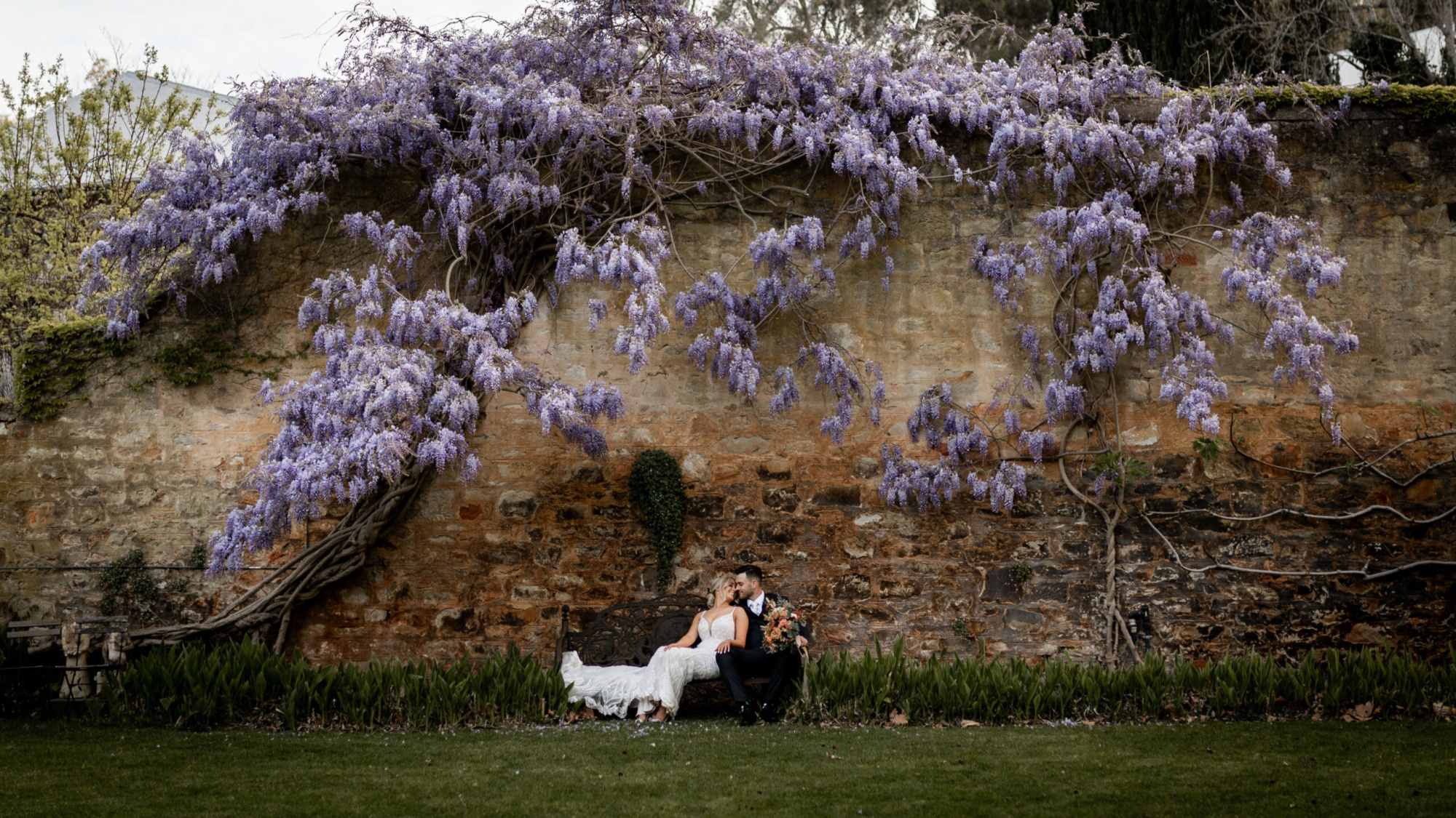 Bride and groom sit on a bench under cascading wisteria against an old stone garden wall.