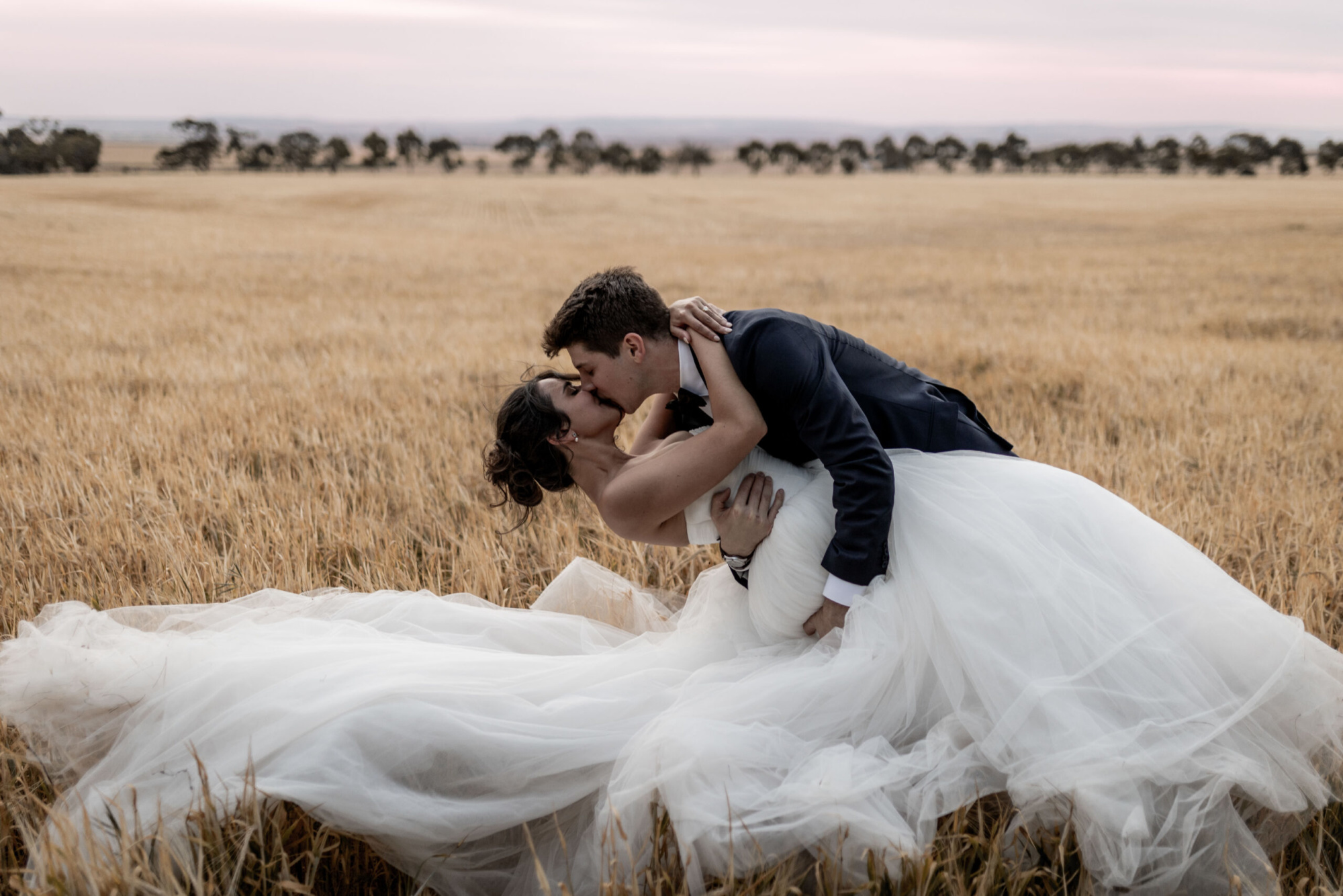 Bride and groom share a romantic kiss as he dips her in a golden field at sunset.