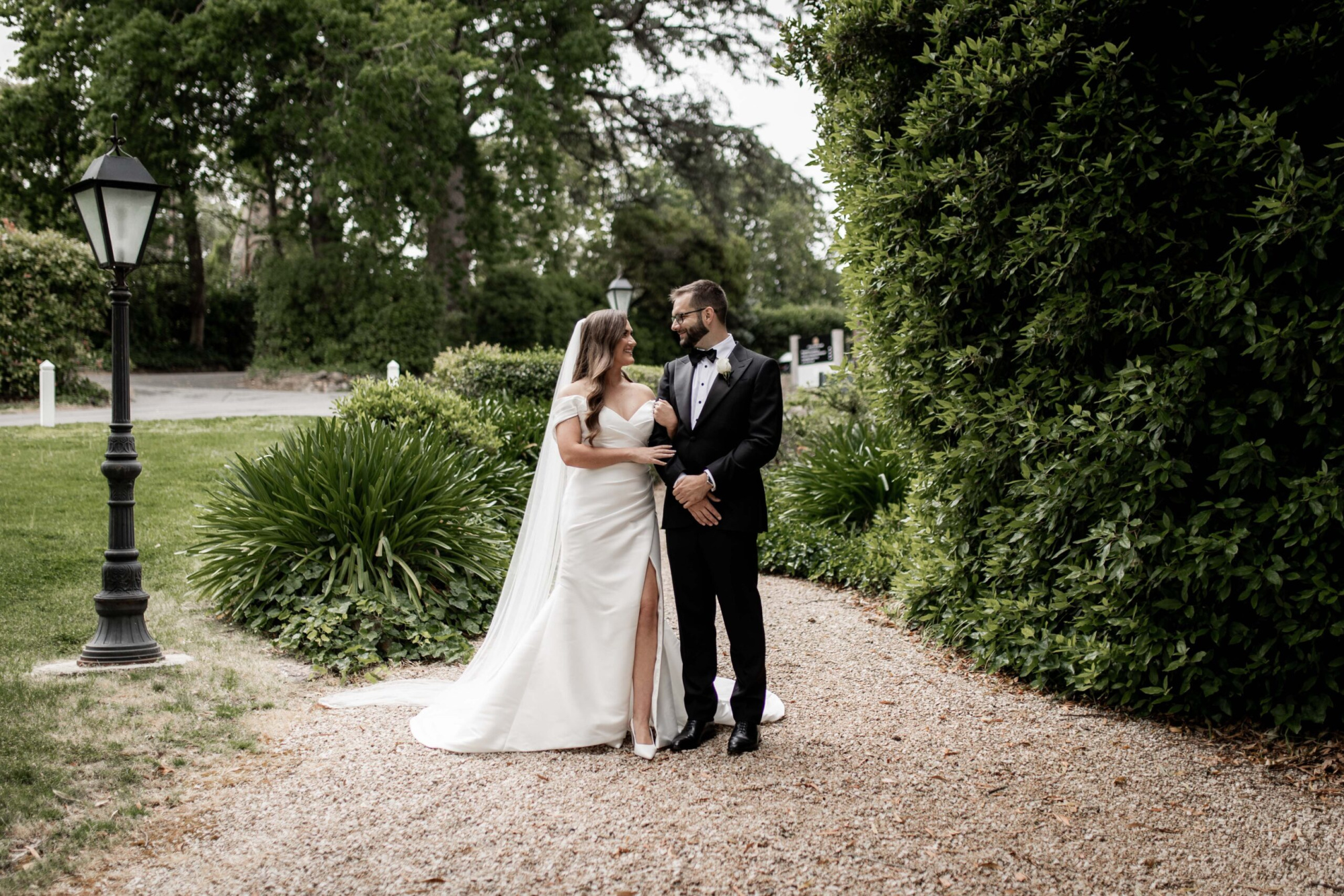 Bride and groom stand on a garden path, smiling at each other amid lush greenery.