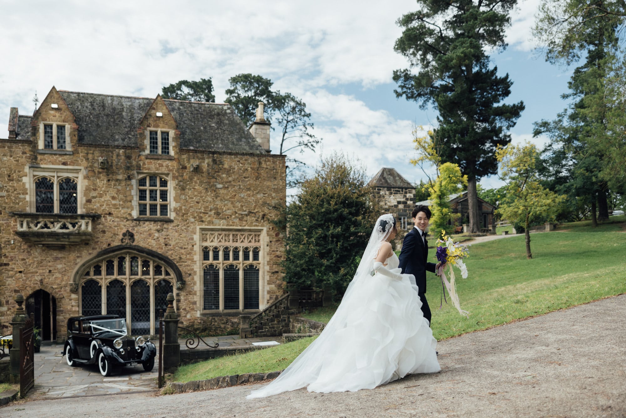 Bride and groom walk outside a grand stone estate with a vintage car parked by the entrance.