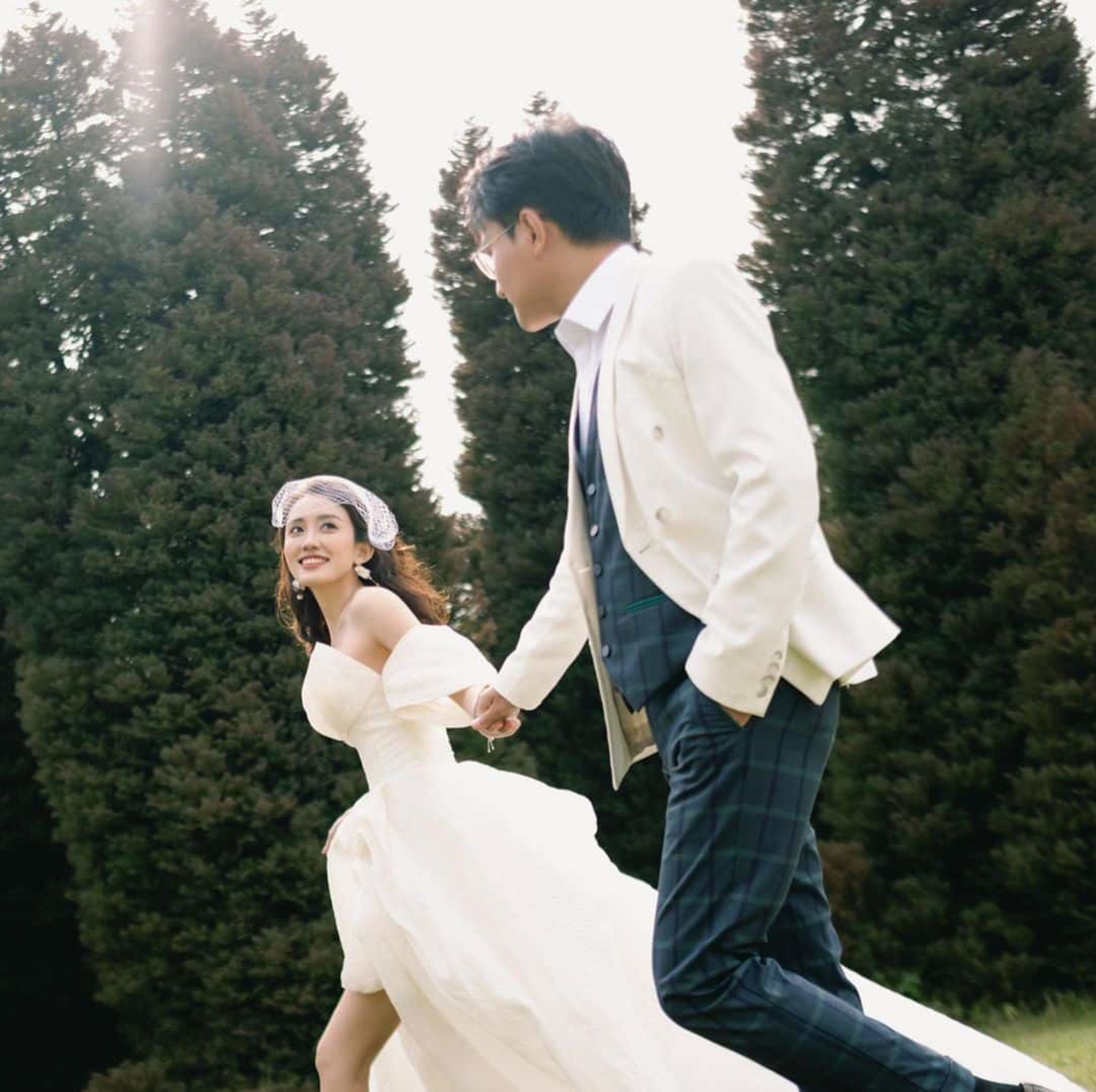 Smiling bride and groom holding hands while running together in a sunlit garden with tall trees behind them.
