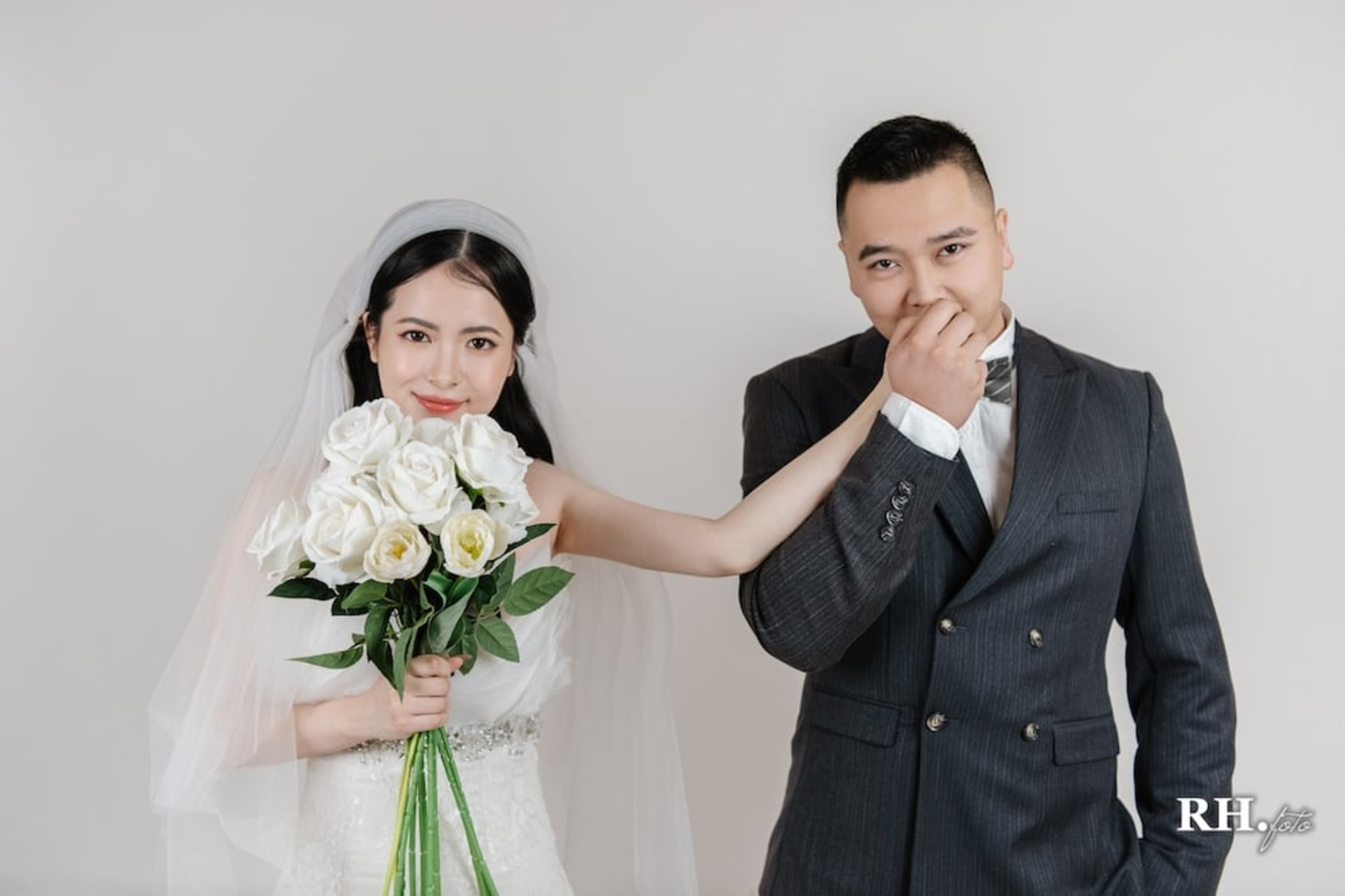 Smiling bride holding white roses as the groom kisses her hand in a studio wedding portrait.