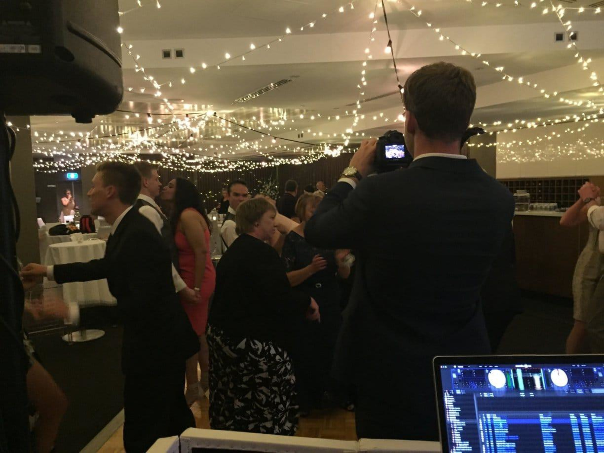 Guests dance under string lights at an indoor wedding reception while a photographer films near the DJ booth.