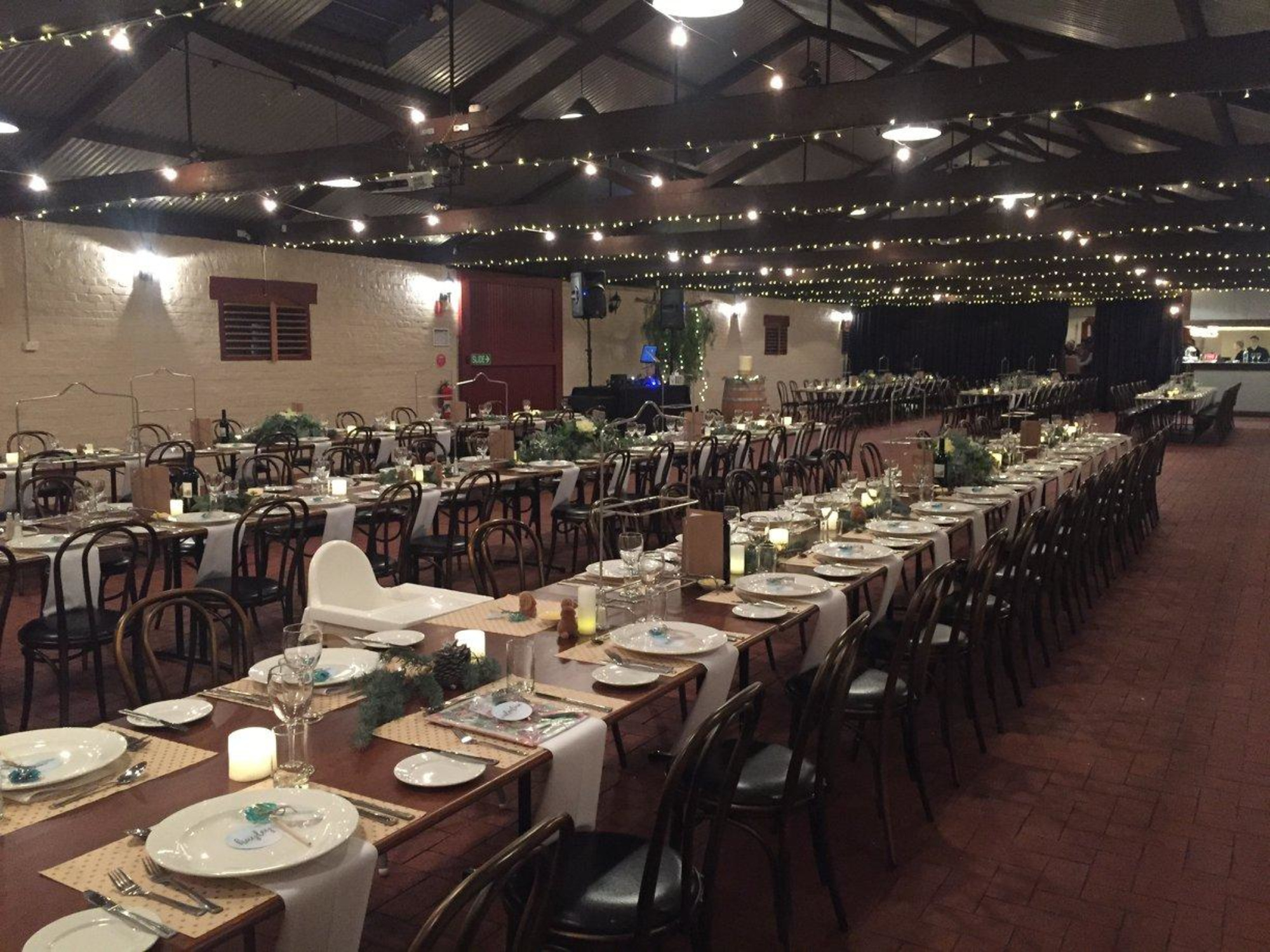 Indoor wedding reception setup with long banquet tables, candles, greenery, and string lights under a high ceiling.