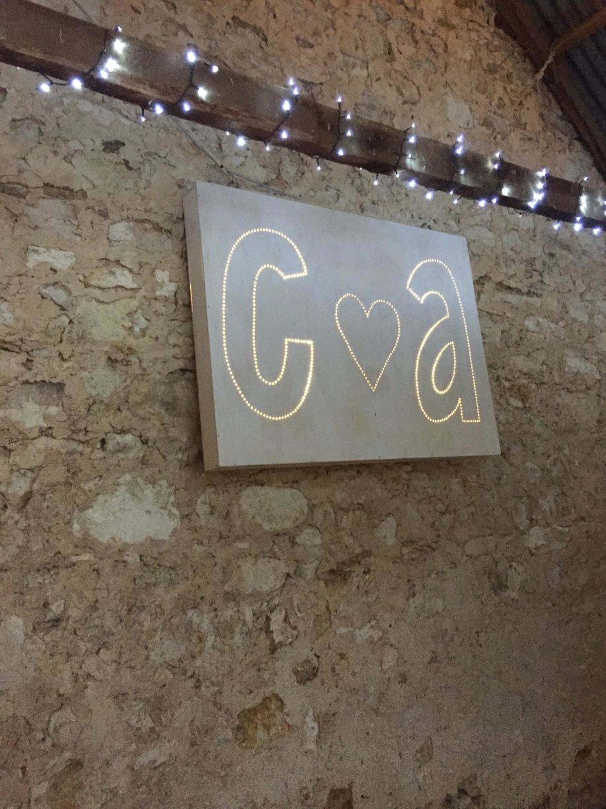 Illuminated initials sign with heart hangs on a rustic stone wall under string lights