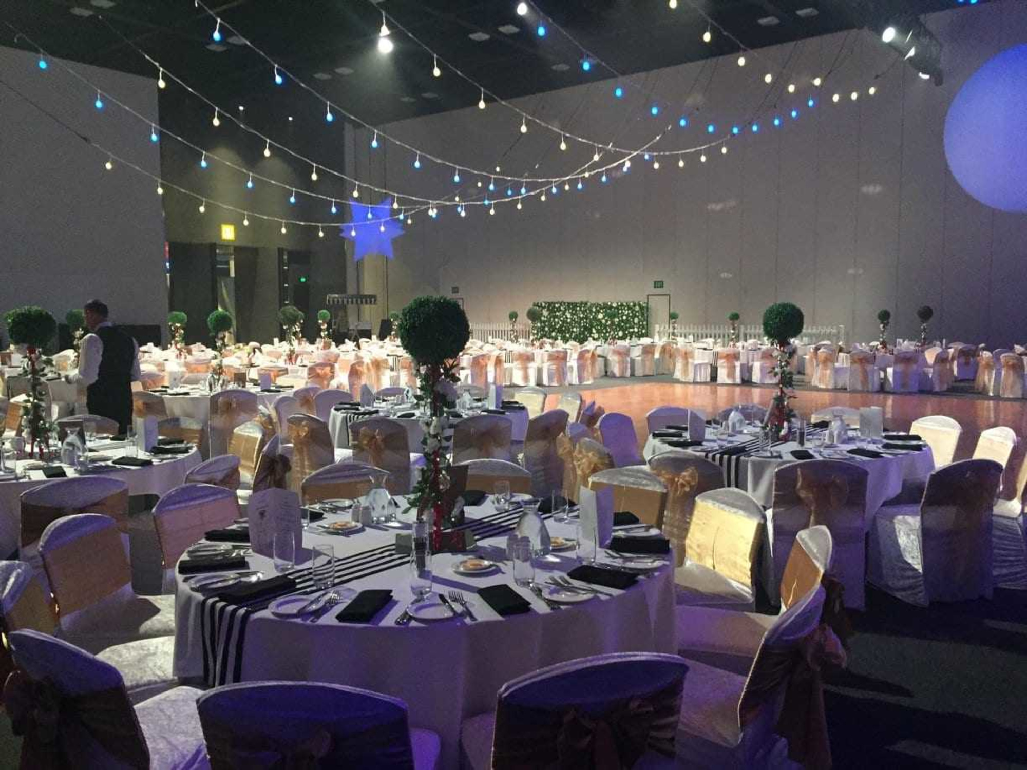 Elegant indoor wedding reception with round tables, gold chair sashes, and string lights over a central dance floor.