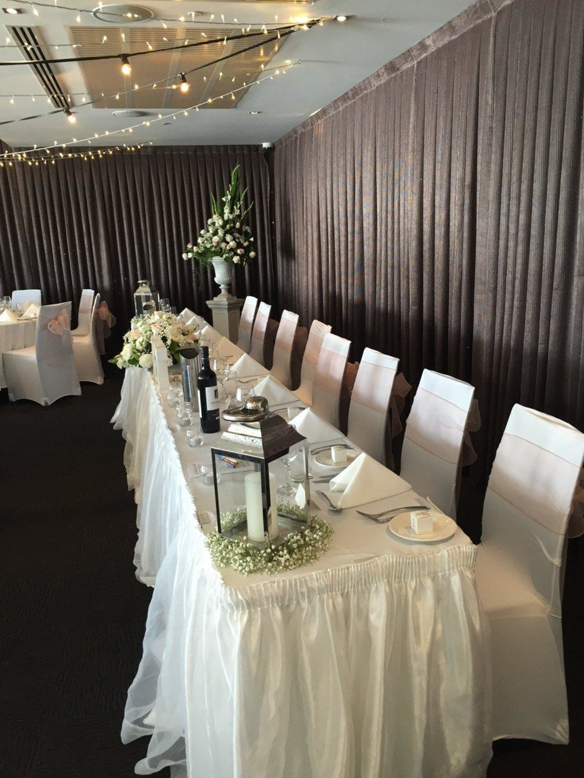 Elegant wedding reception head table with white linens, lantern centerpieces, and fairy lights overhead.