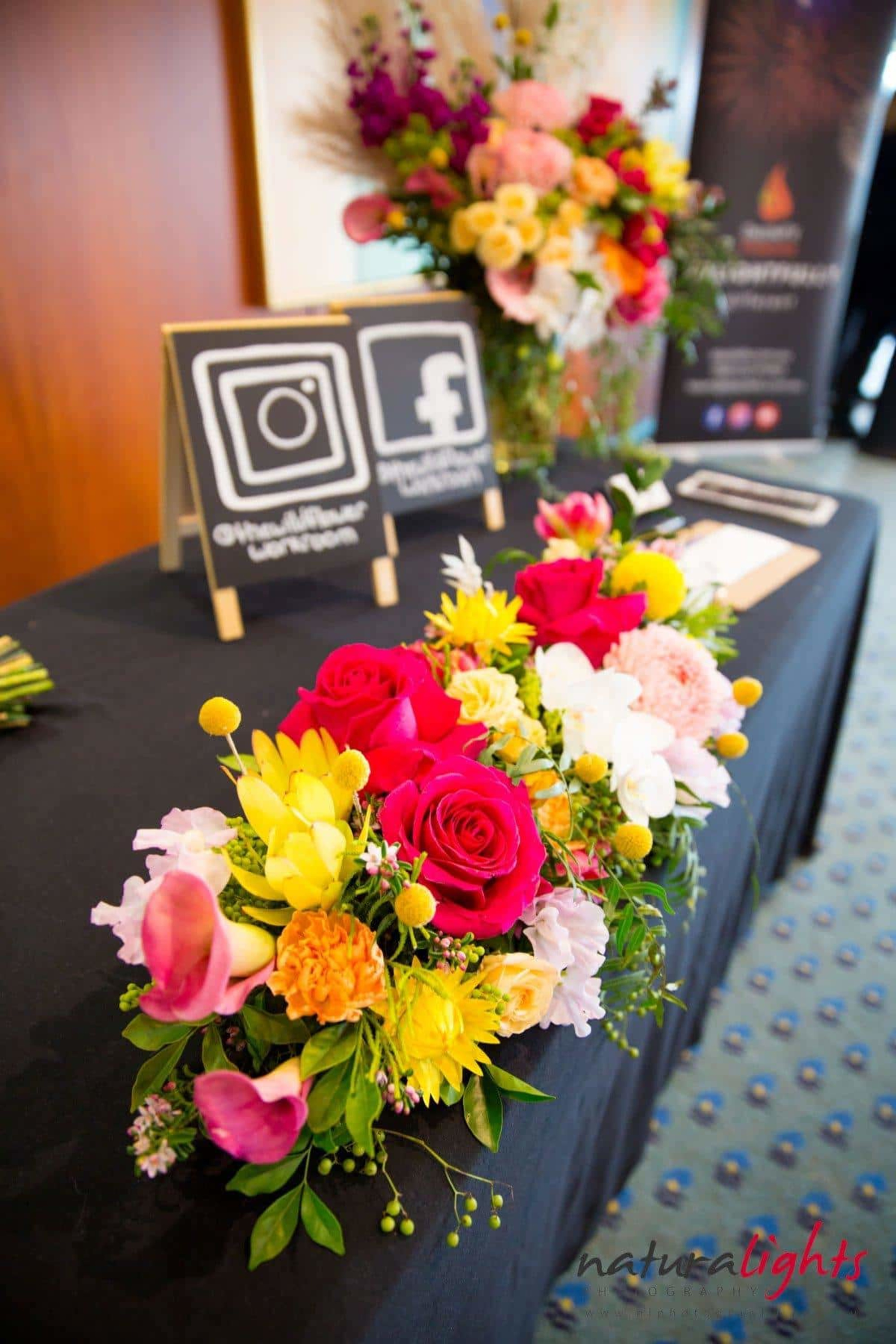Colorful floral centerpiece on a black-covered vendor table at a wedding showcase with social media signs in the background.