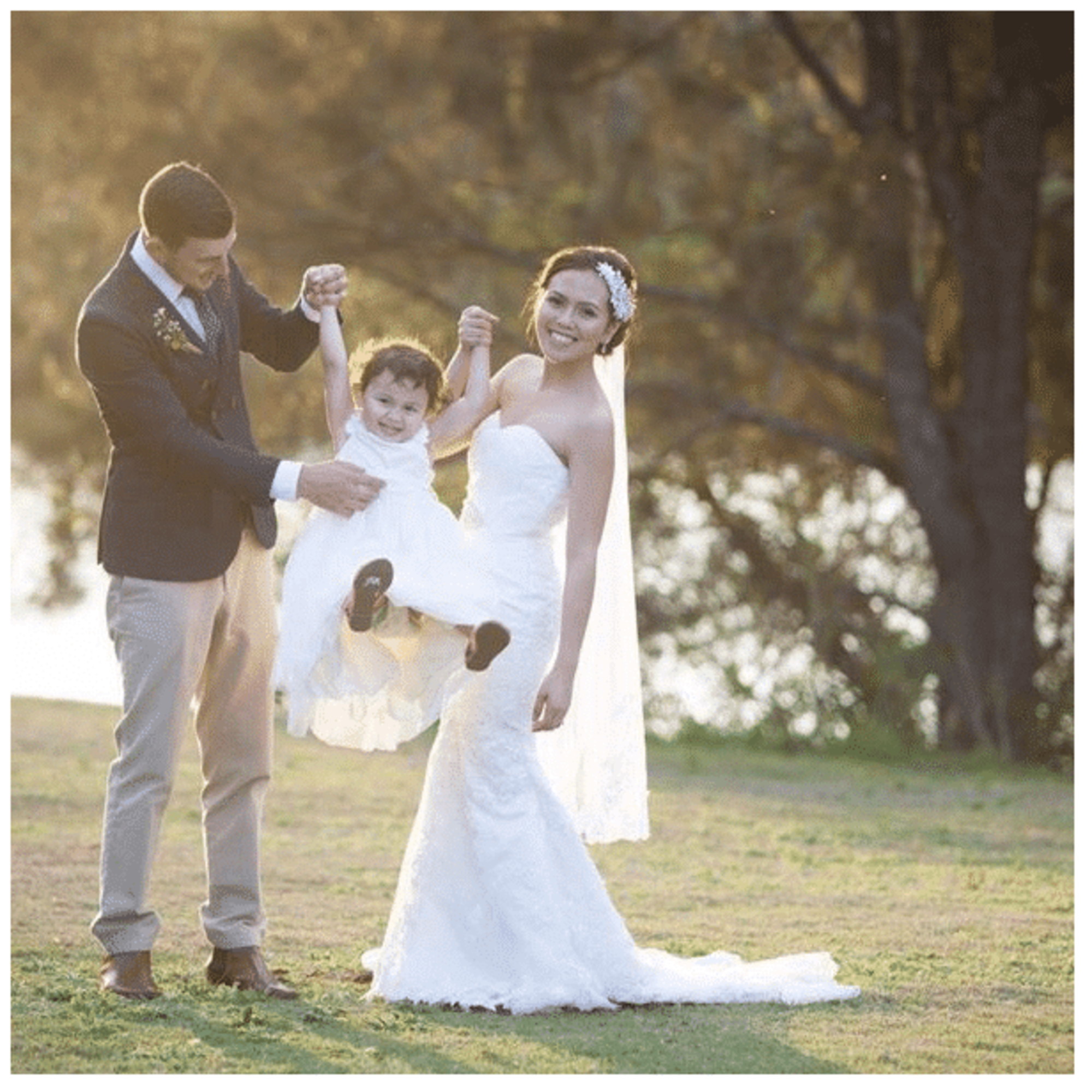 Bride and groom swing a smiling child between them during an outdoor wedding at sunset.