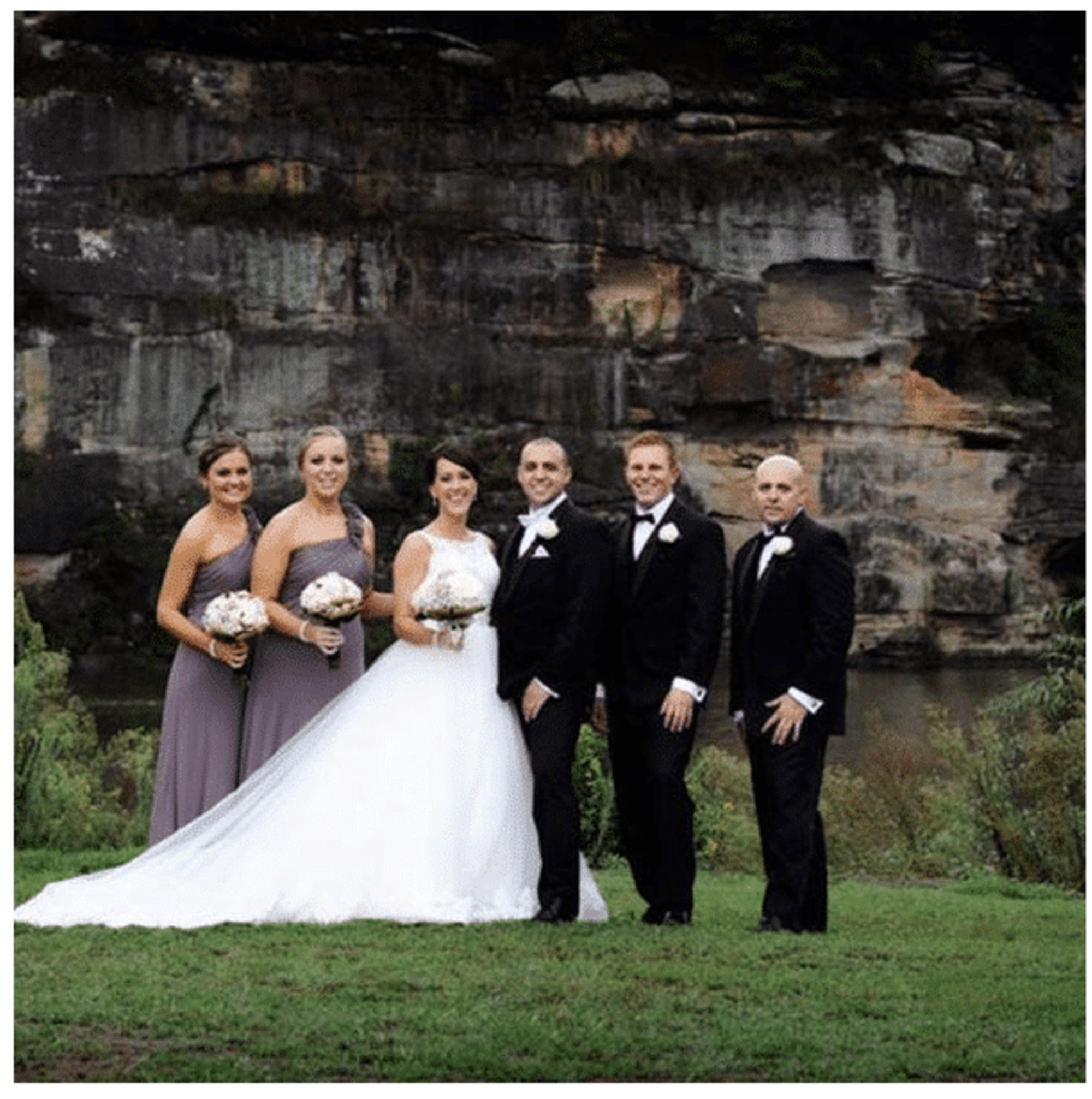 Formal wedding party posing outdoors in front of a rocky cliff and greenery.