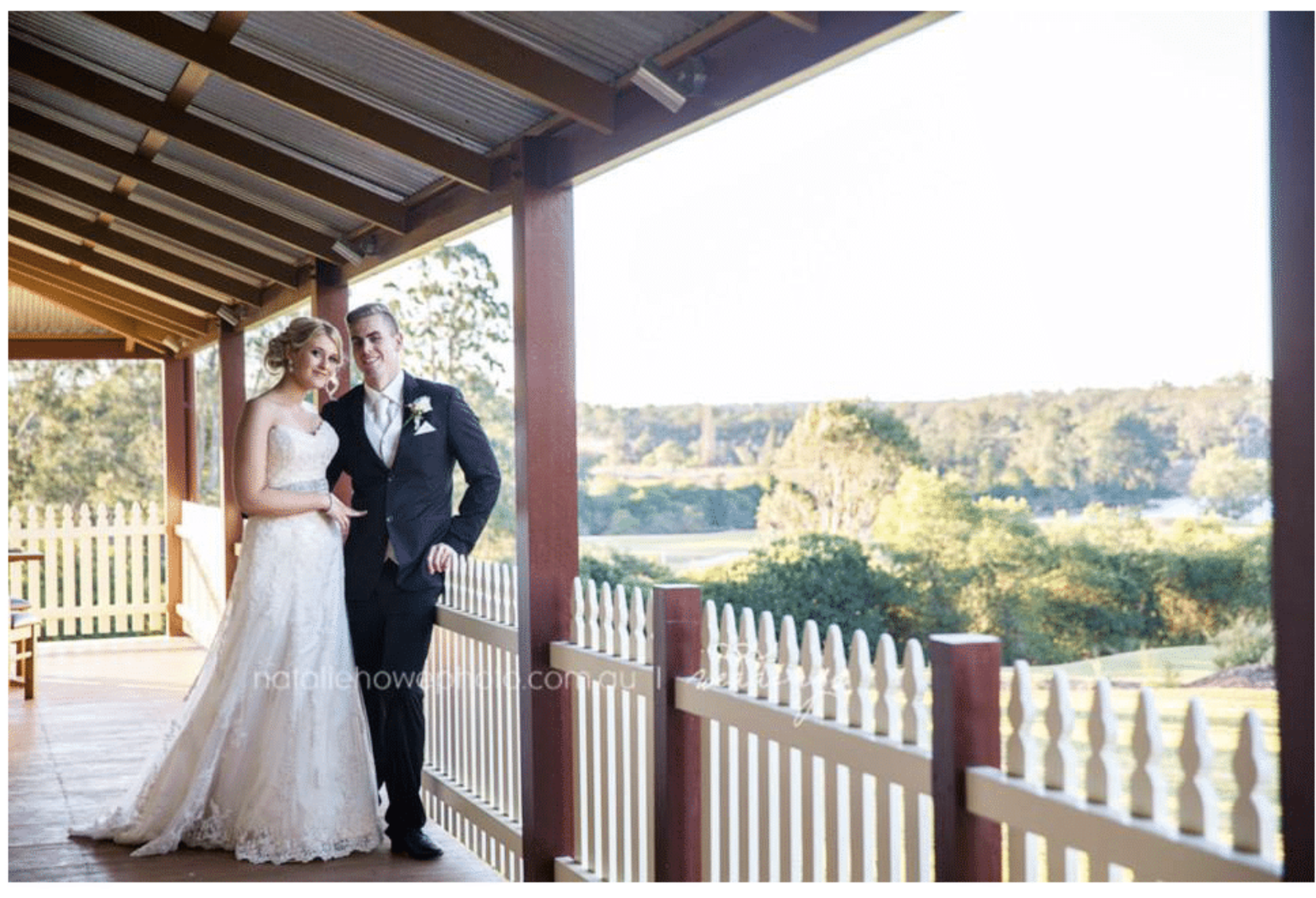 Bride and groom pose on a sunny country veranda overlooking green fields and trees.