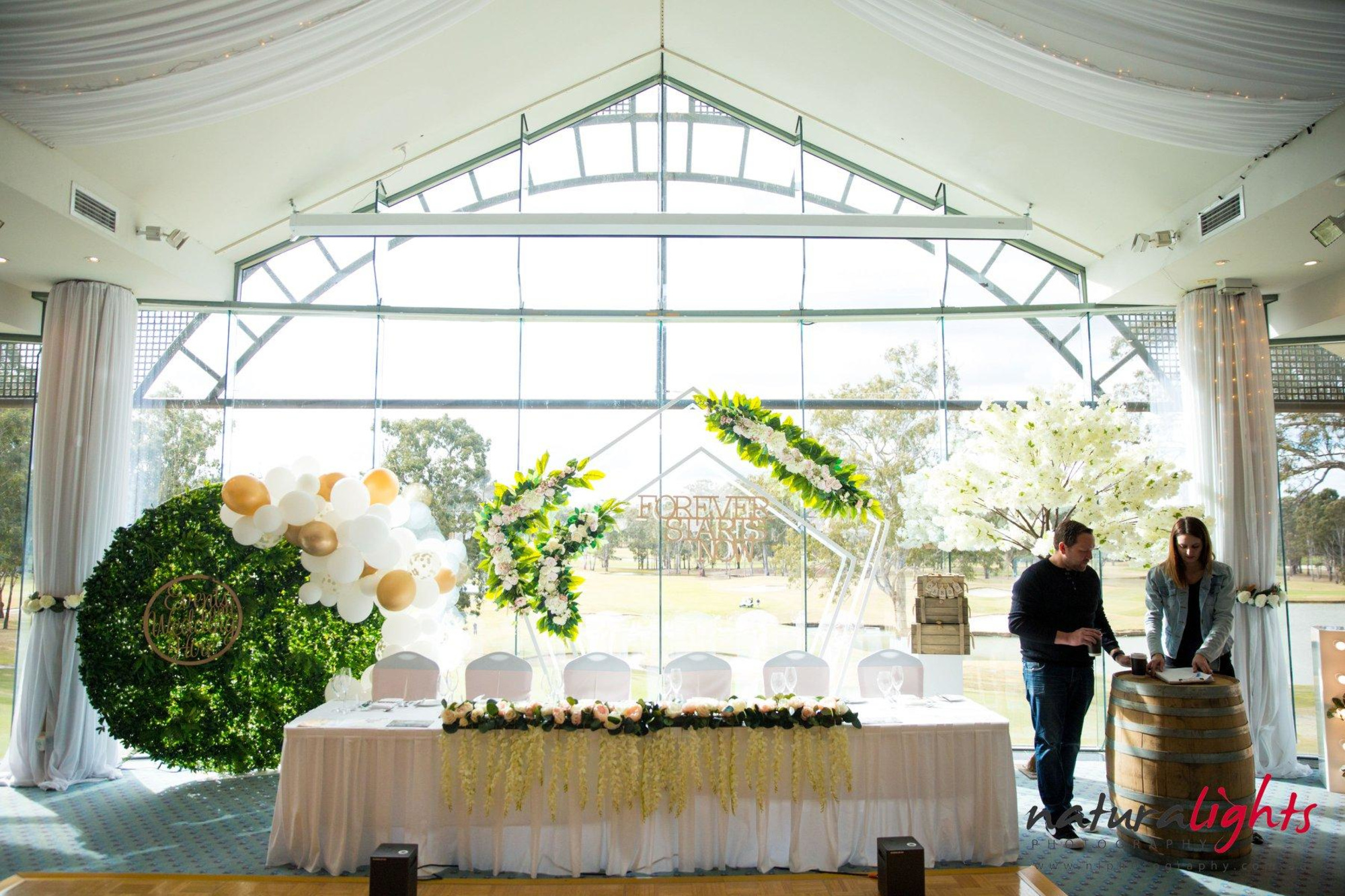 Bright indoor wedding reception setup with floral backdrop, balloon garland, and decorated head table by large windows.