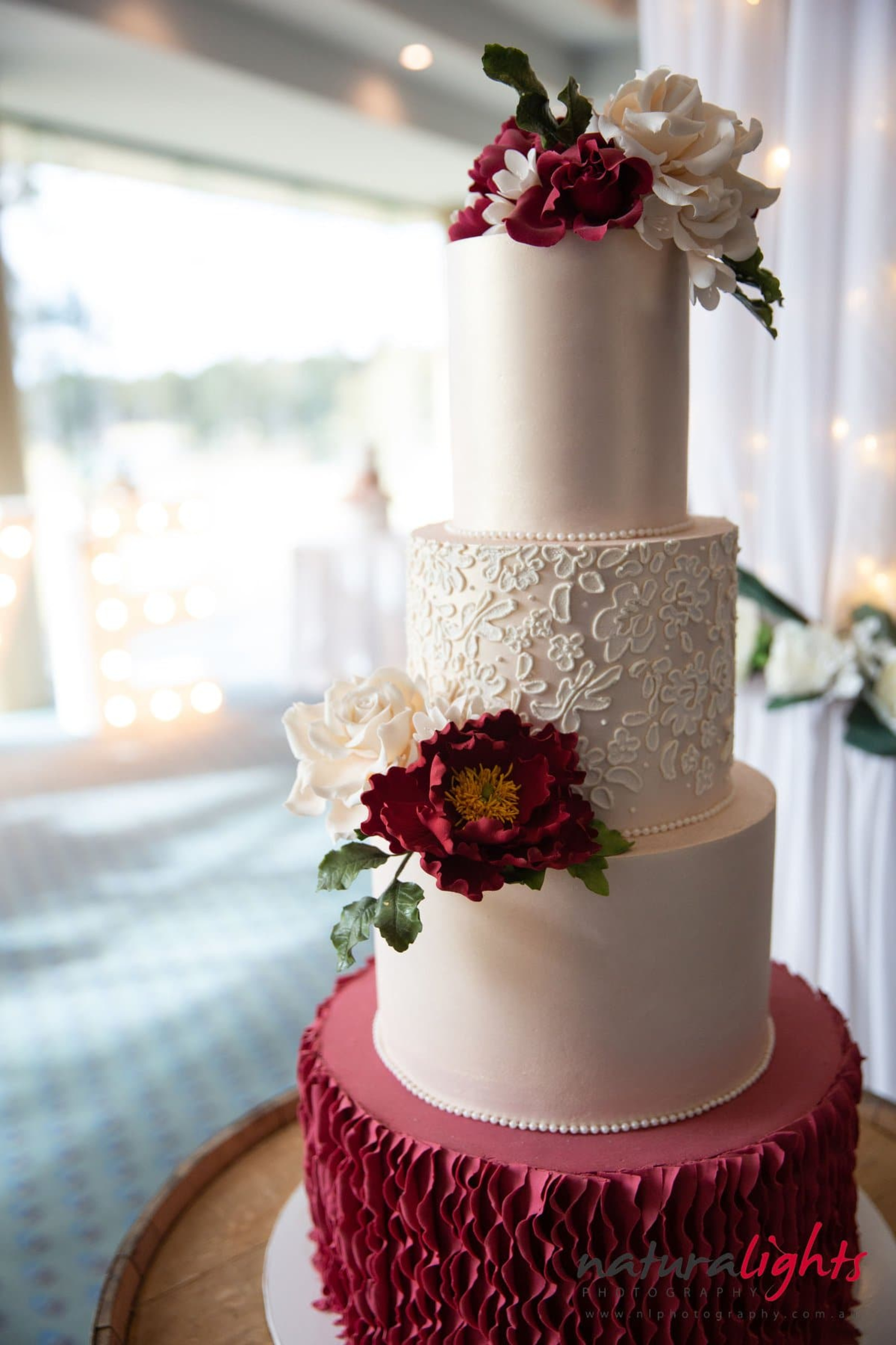 Three-tier ivory and burgundy wedding cake with sugar flowers and lace detailing on a wooden stand.