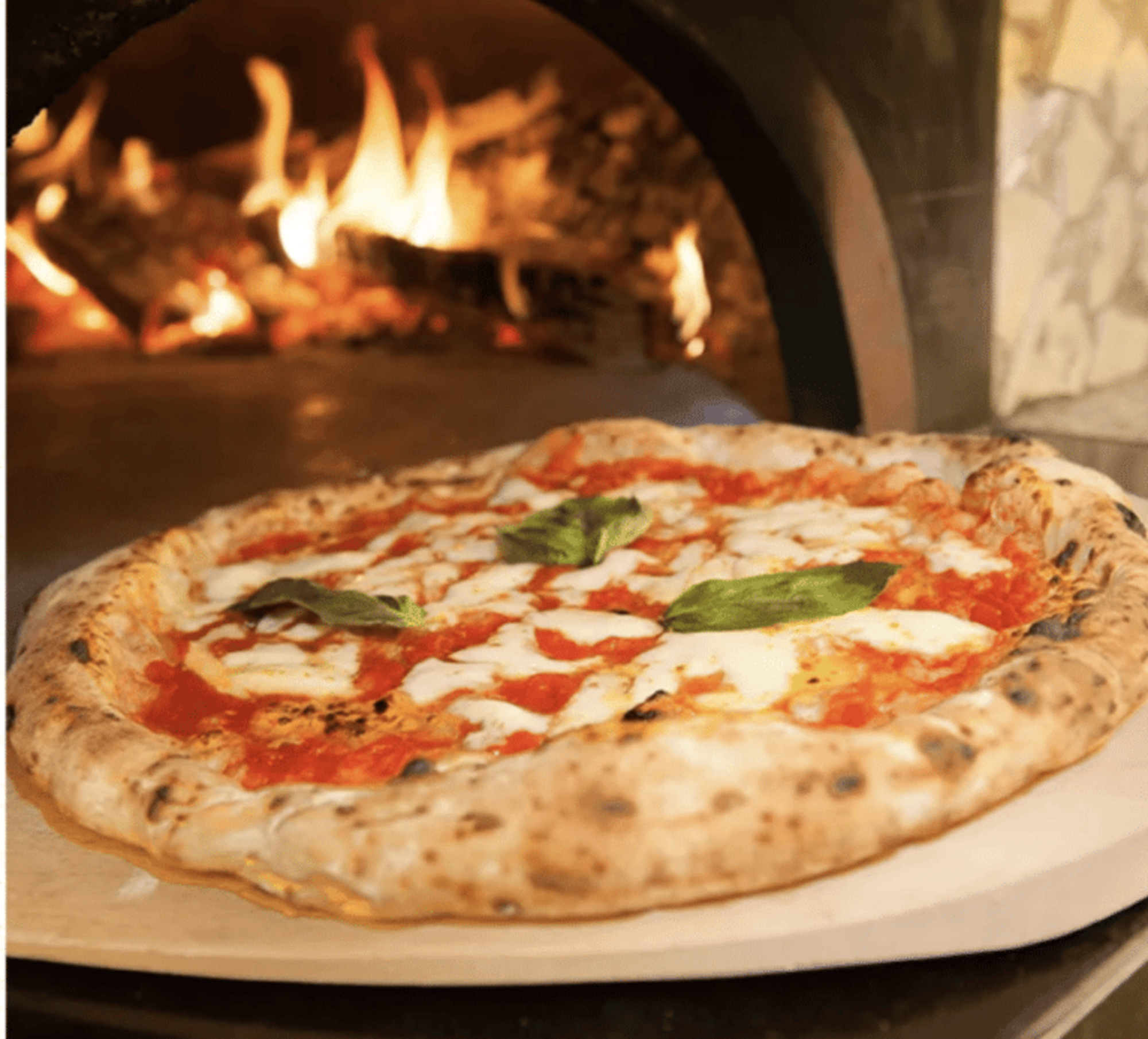 Wood-fired margherita pizza baking in a stone oven with flames in the background.