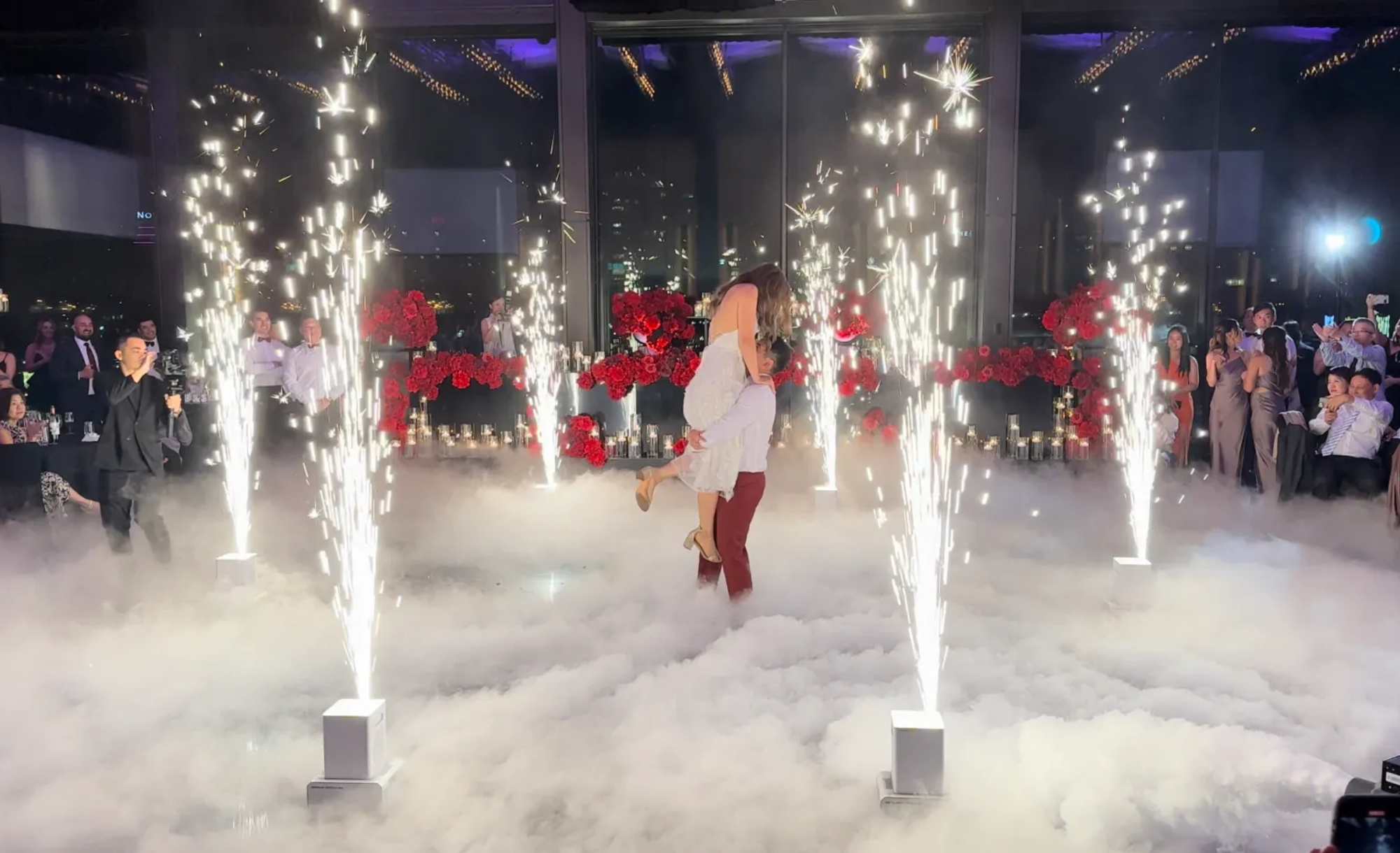 Couple dances amid fog and indoor sparkler fountains at a red-rose filled wedding reception.